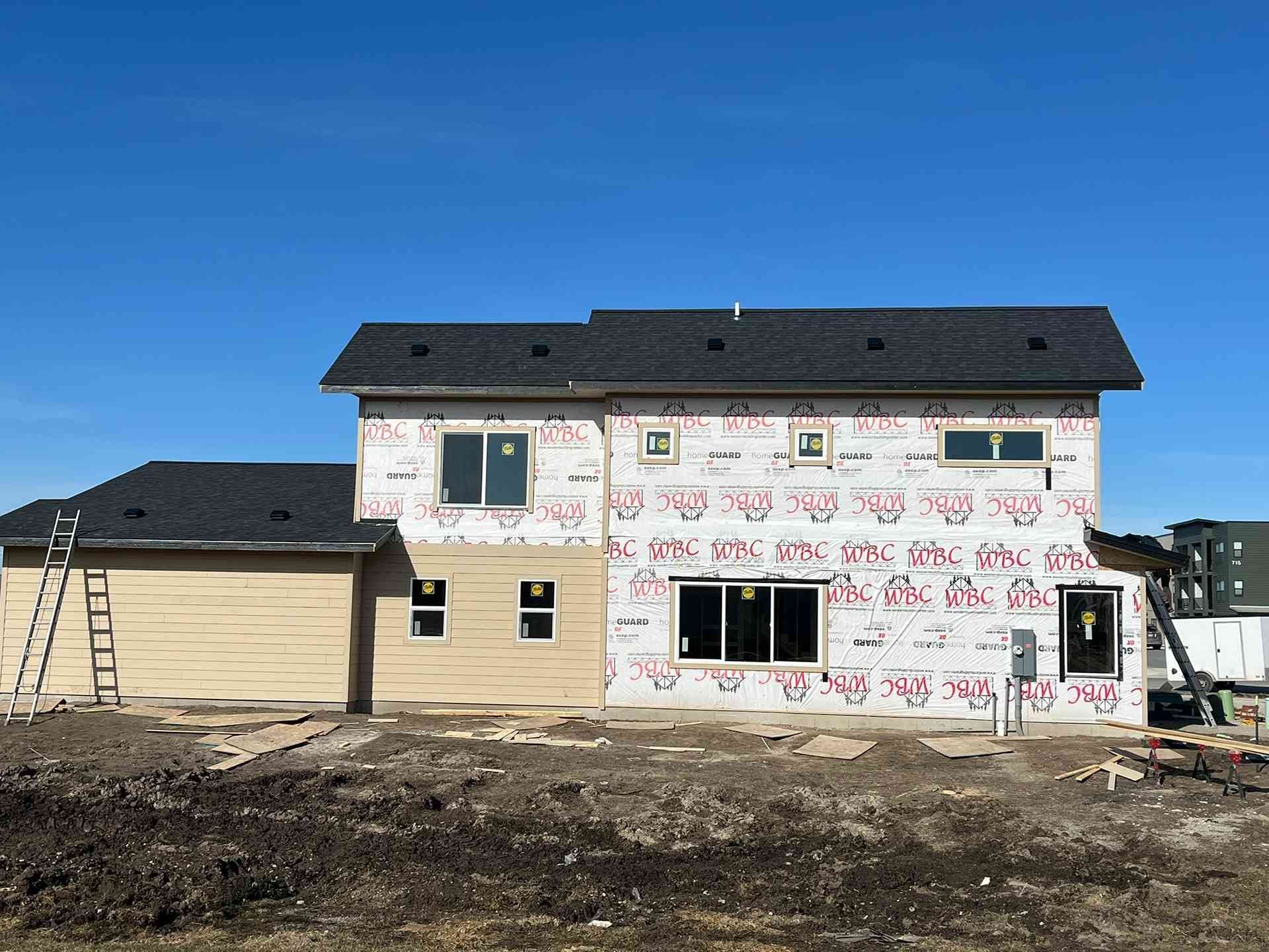 Partially built two-story house with white wrap, black roof, and ladders on dirt lot under blue sky