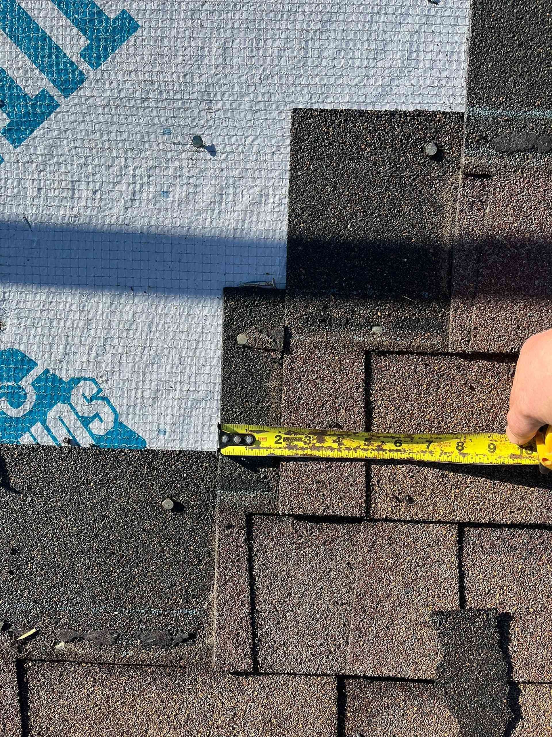 Tape measure over roof shingles at a seam between gray underlayment and brown shingles