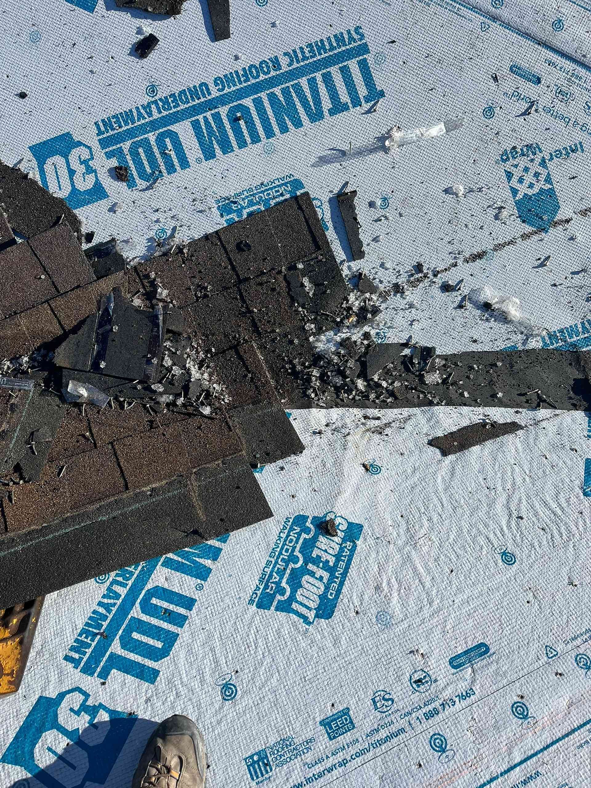 Close-up of a damaged roof with torn black shingles over blue-and-white underlayment.