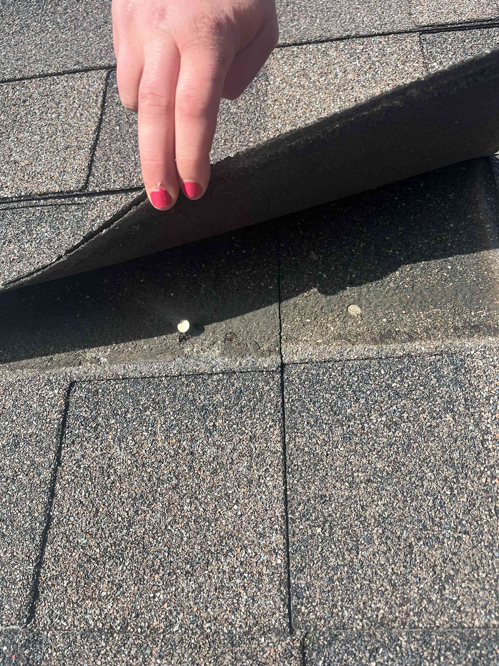 Hand lifting a curled shingle on a gray asphalt roof, exposing the seam beneath