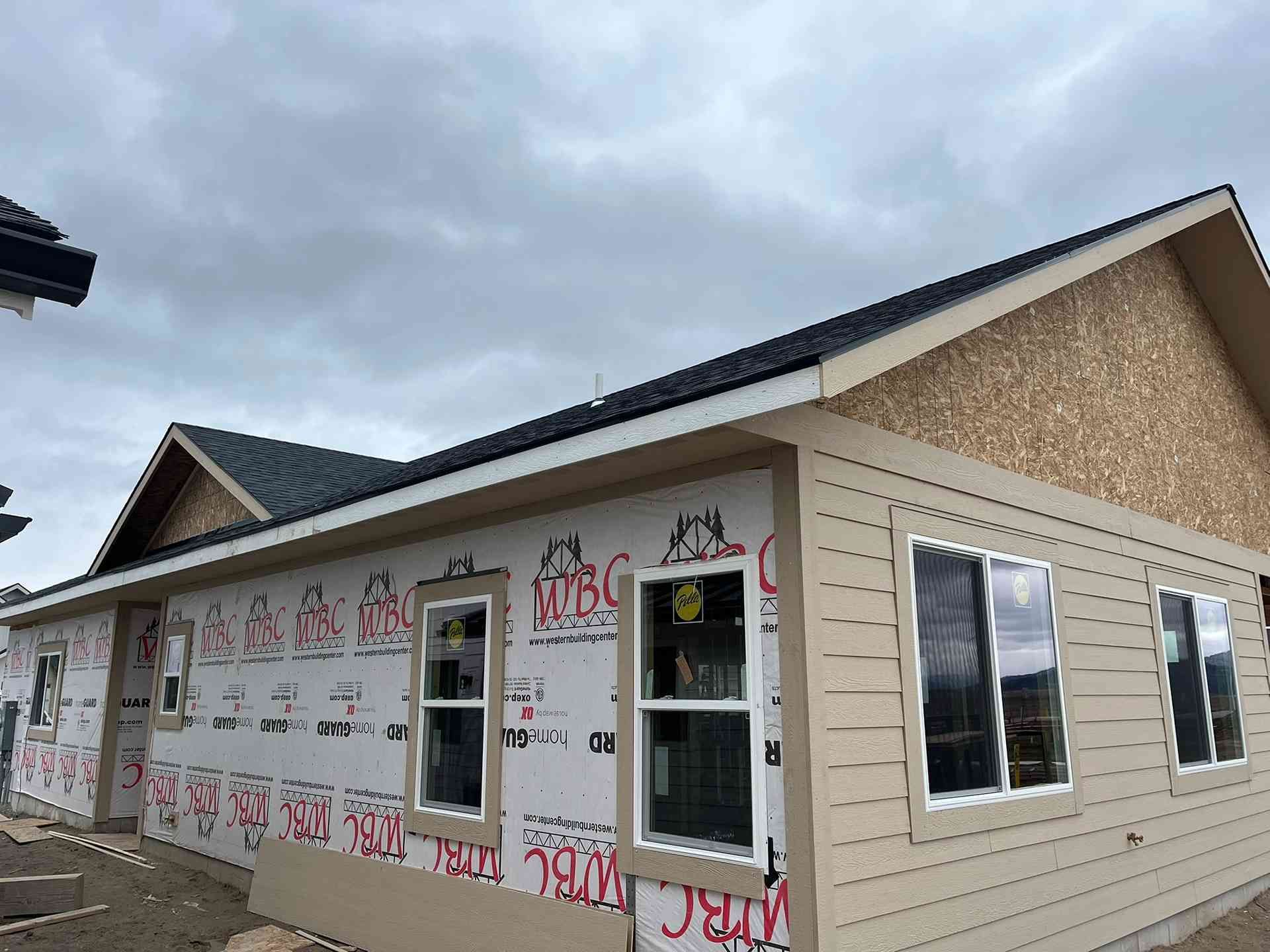 Partially built beige house with wrapped exterior, windows, and dark roof under cloudy sky