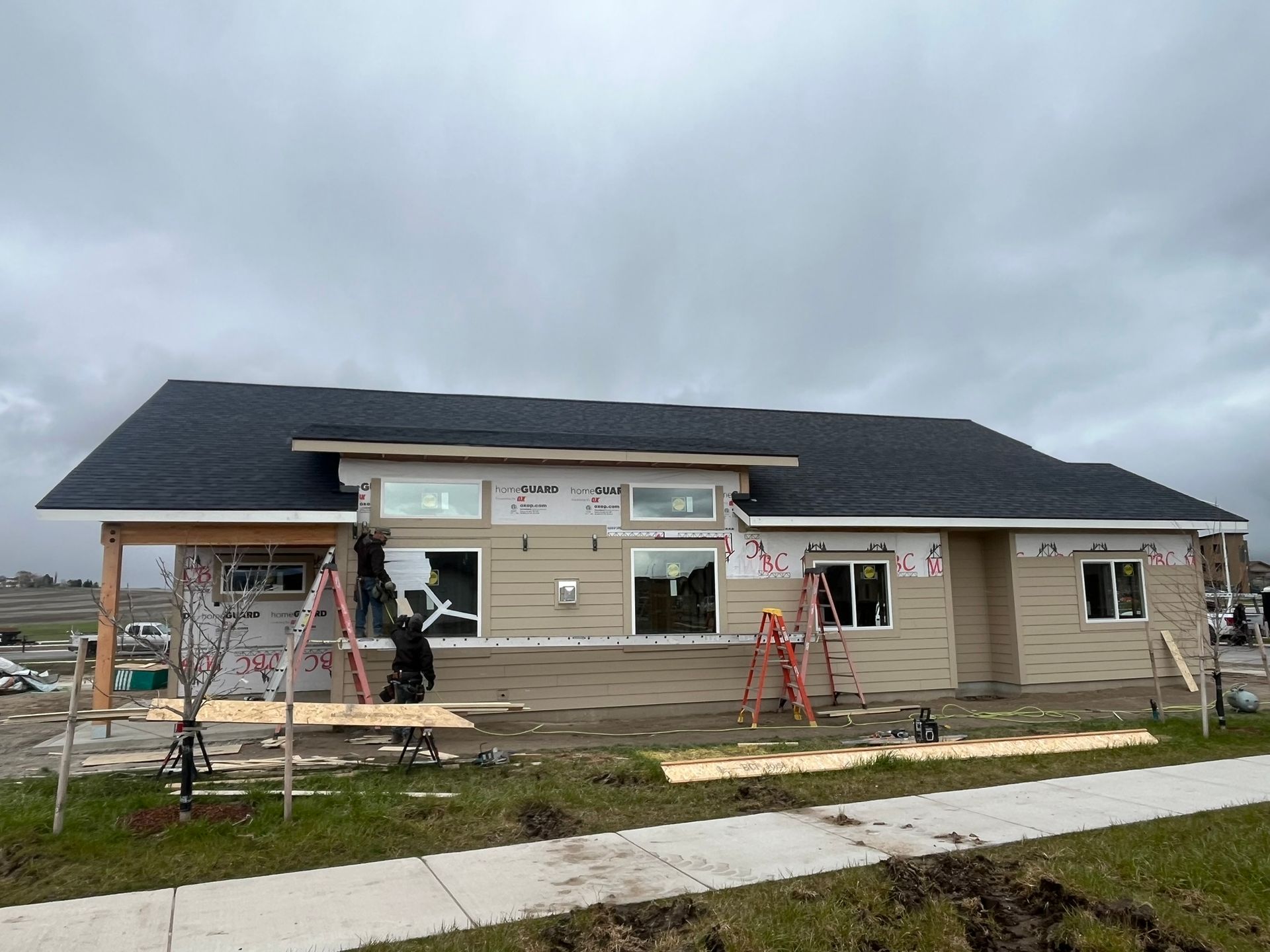 House under construction with workers, ladders, and exposed siding beneath a cloudy sky