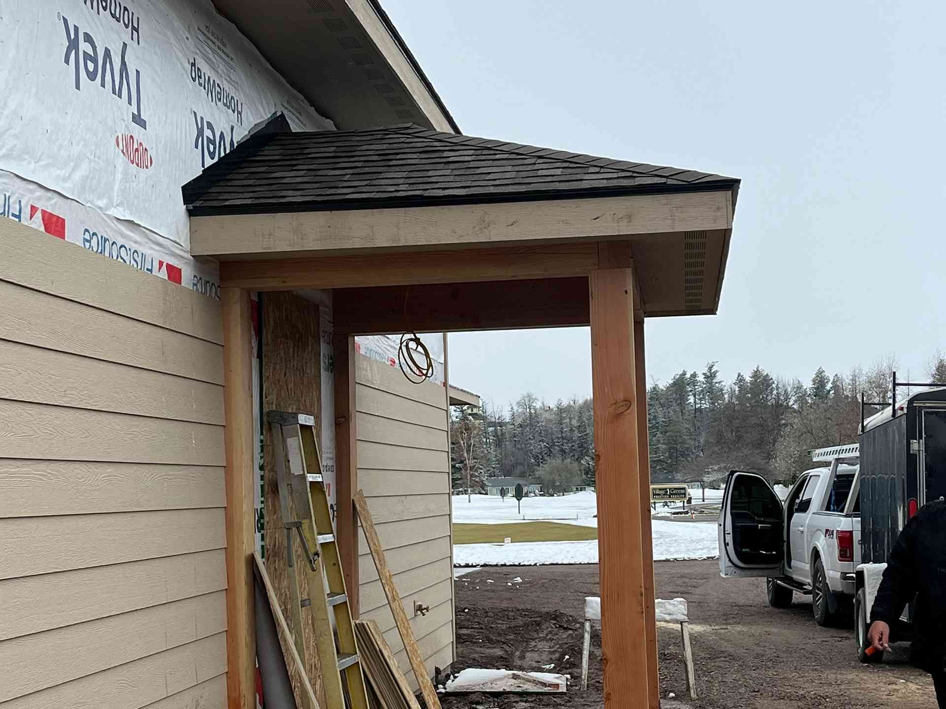 Covered wooden entryway beside a beige building, with a van parked on a snowy roadside.