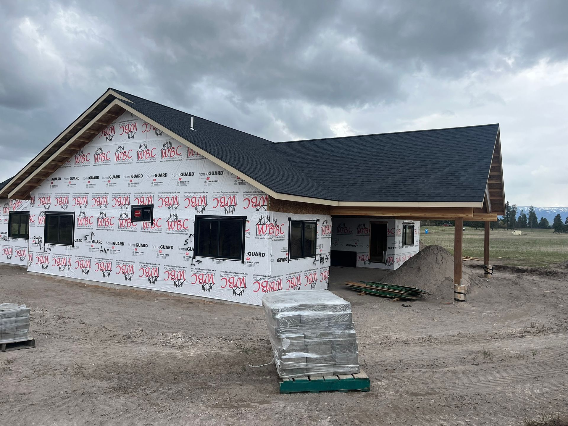 Partially built house with white wrap and dark roof on a gravel lot under cloudy skies