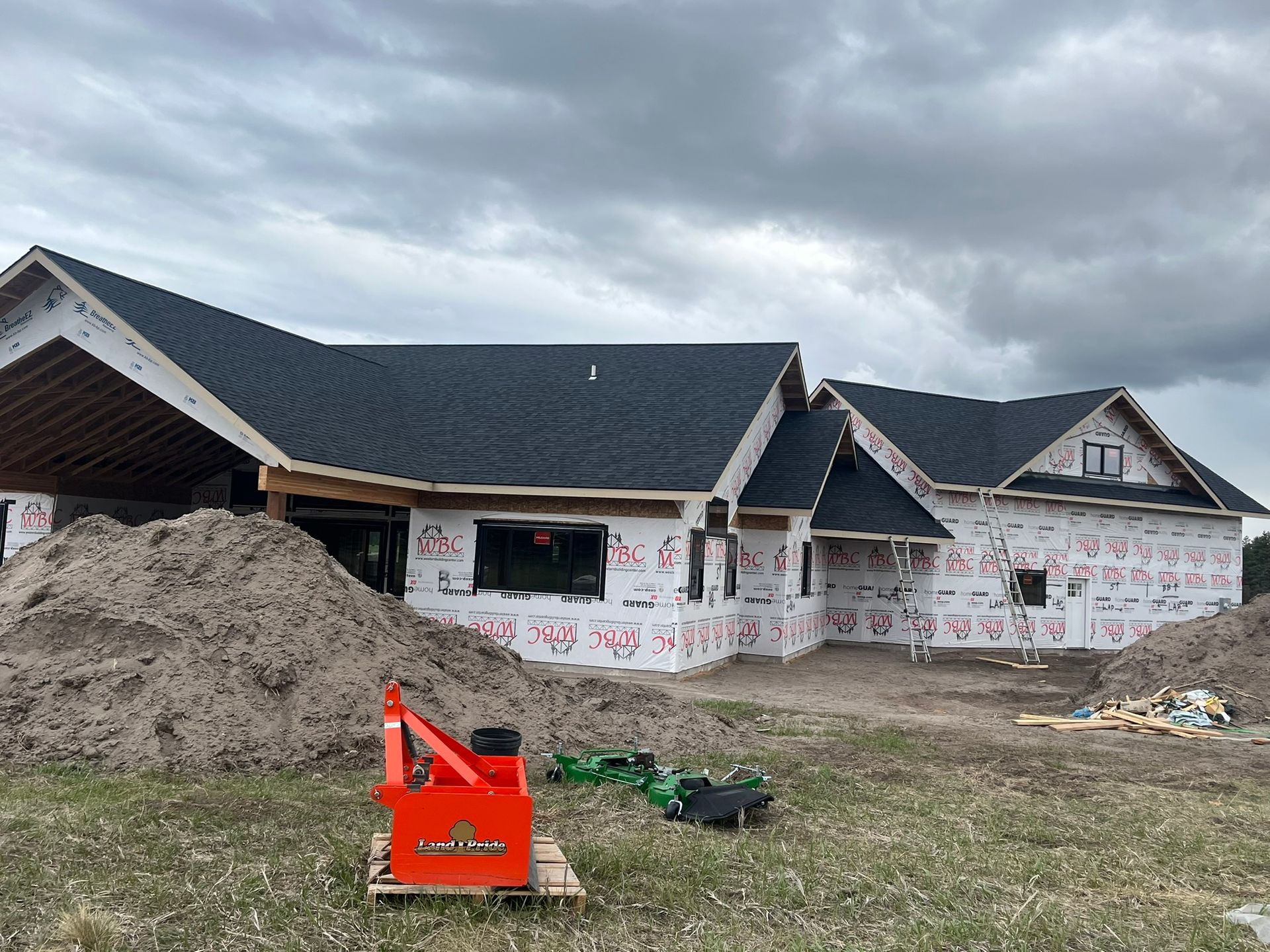 Construction site with a partially built house, large dirt pile, and orange equipment under cloudy skies