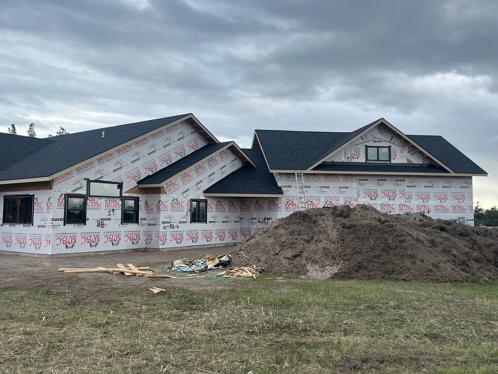 Unfinished house under cloudy sky, wrapped in white insulation with a large dirt pile in front.