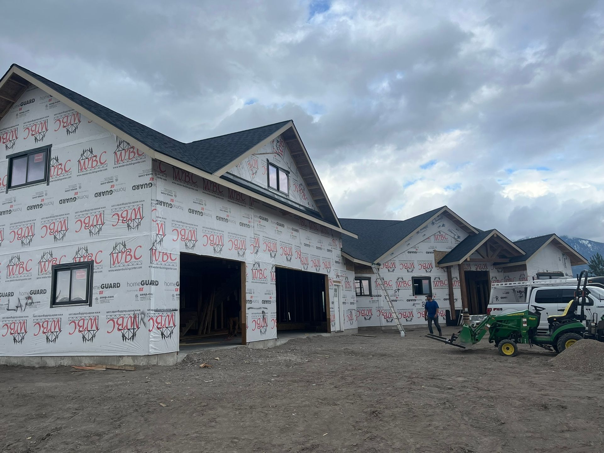 Partially built house wrapped in white housewrap, with open garage bays and tractors on a gravel lot