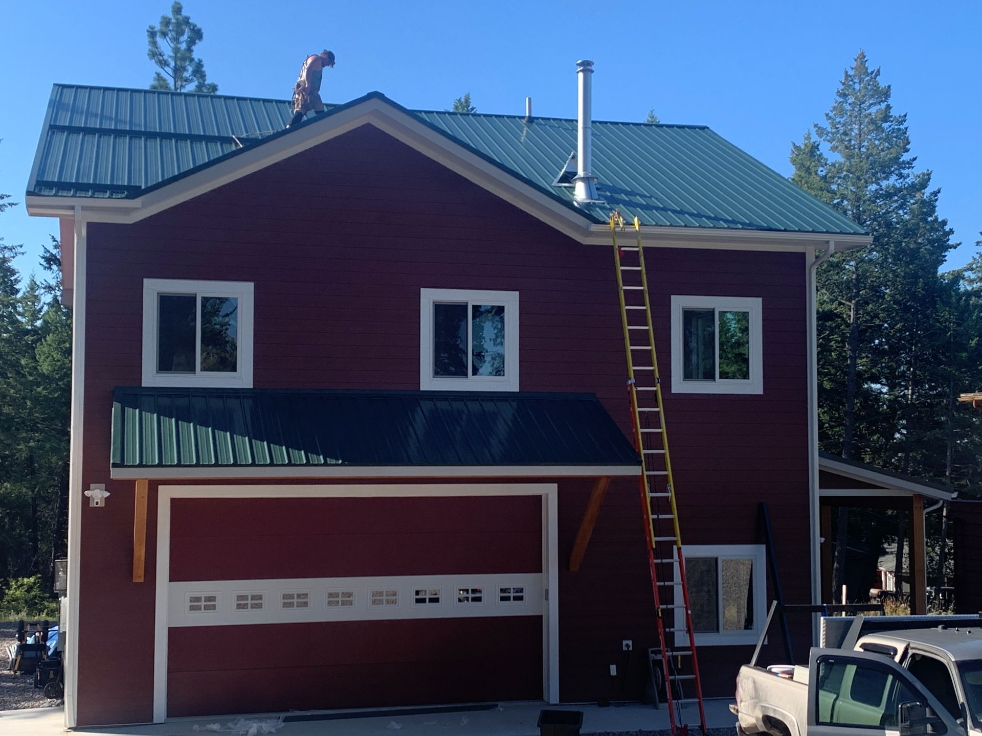 Red house with green metal roof, garage door, and ladder leaning against the front under a blue sky