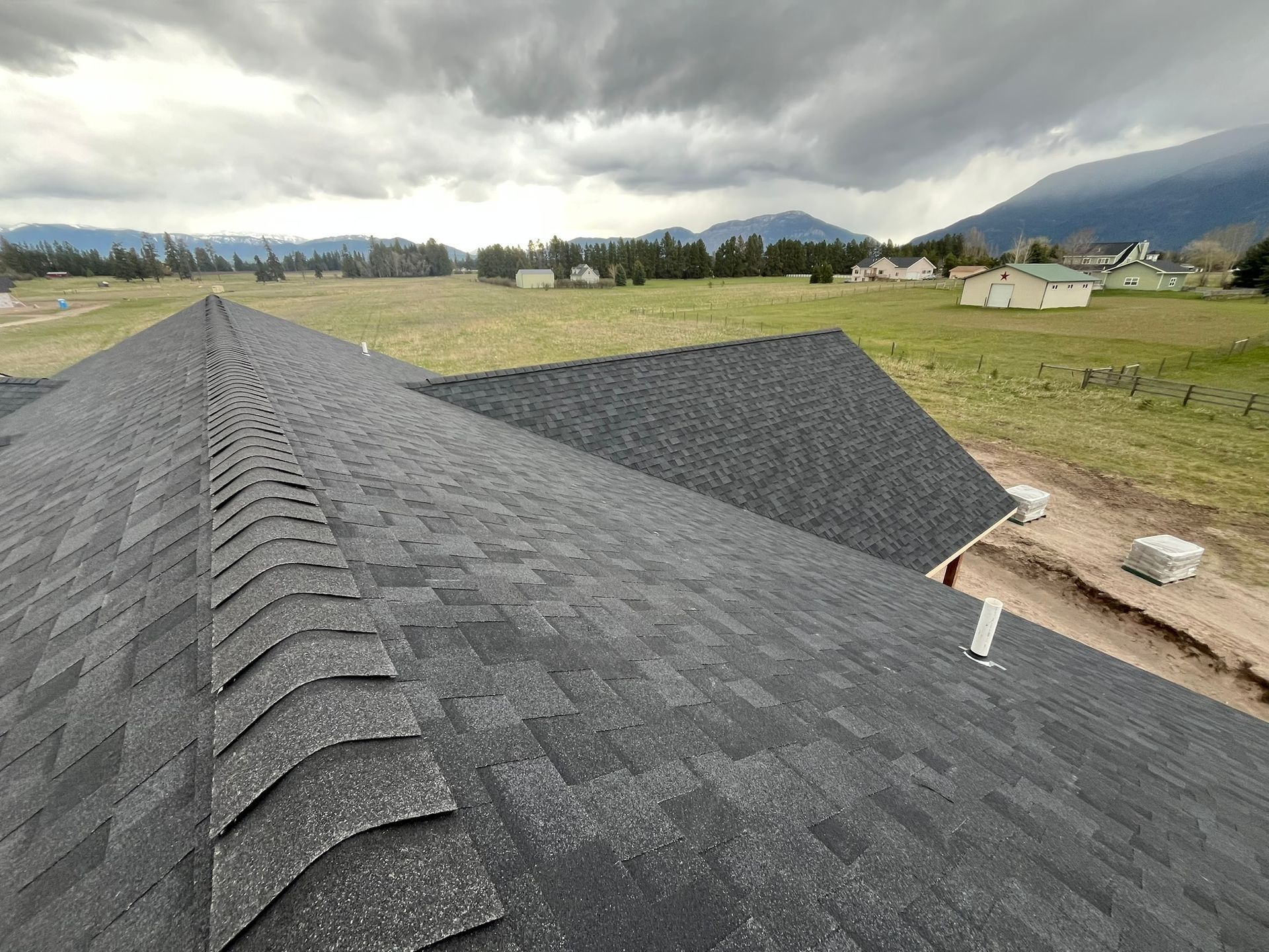 Dark shingled roof in foreground overlooking a rural field and mountains under cloudy skies