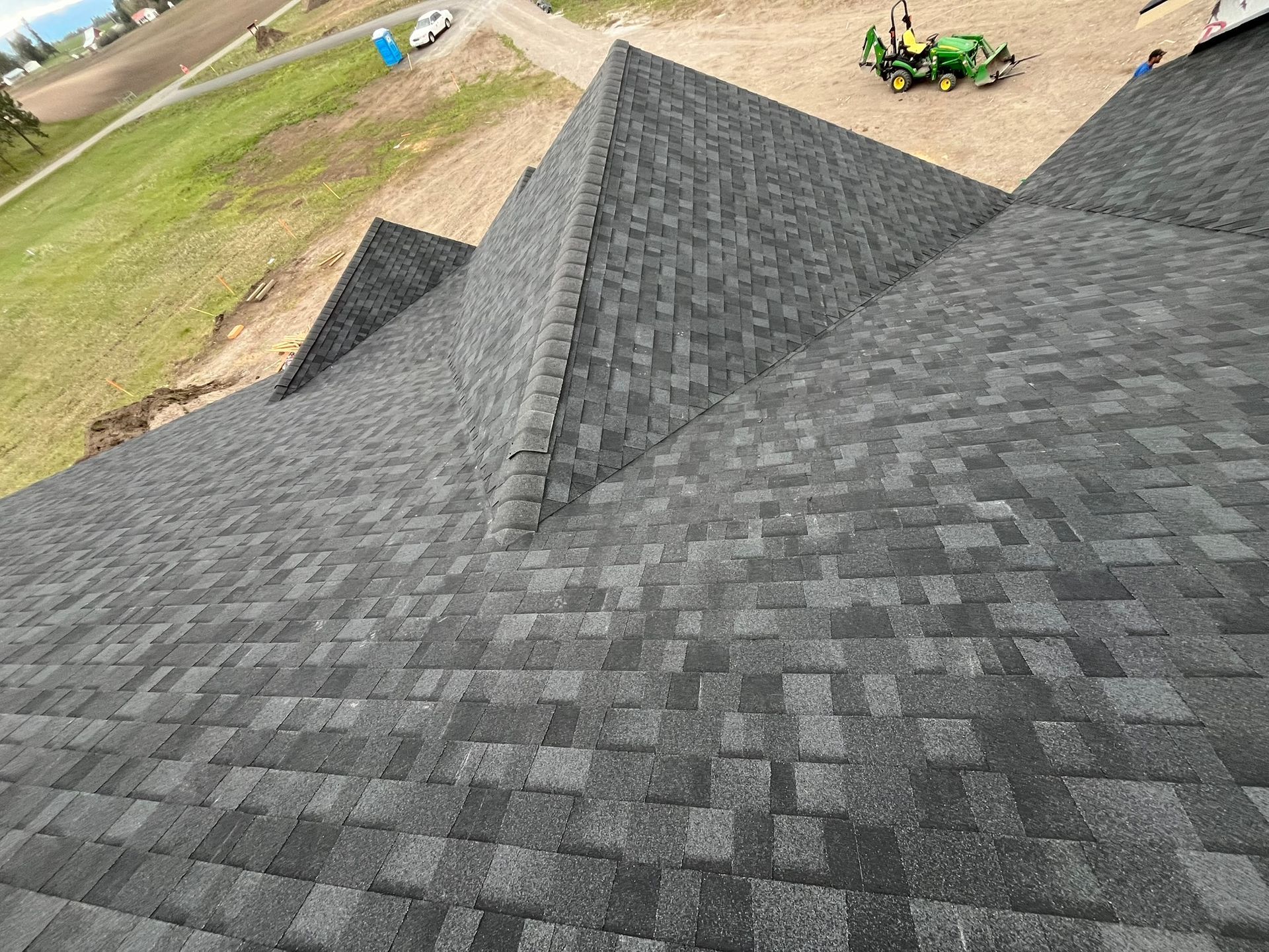 Gray shingle rooftops on a house under construction, viewed from above, with a green tractor in the background