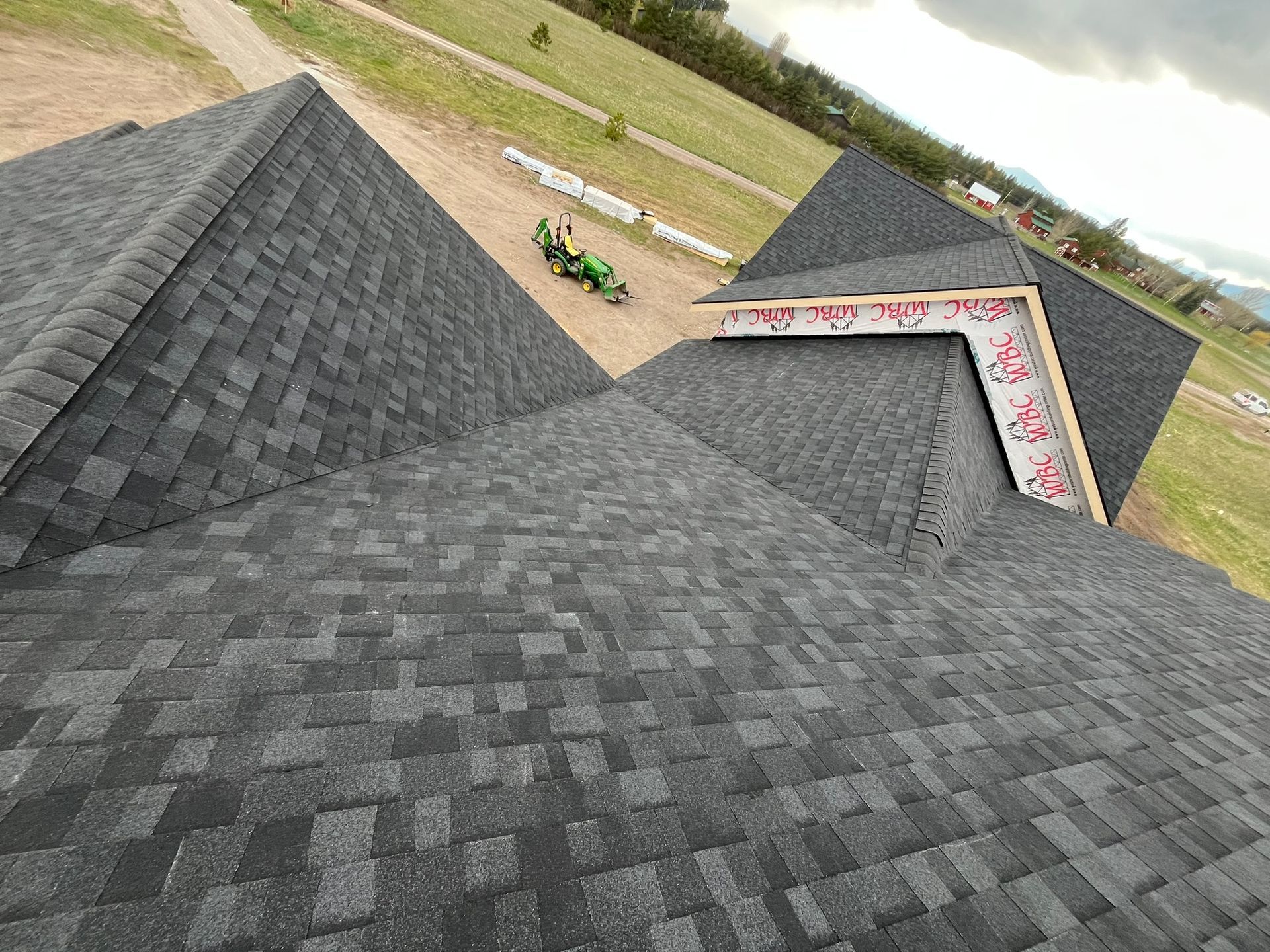 View from a roof ridge over dark gray shingles toward a road, grassy area, and a green tractor near a small building.