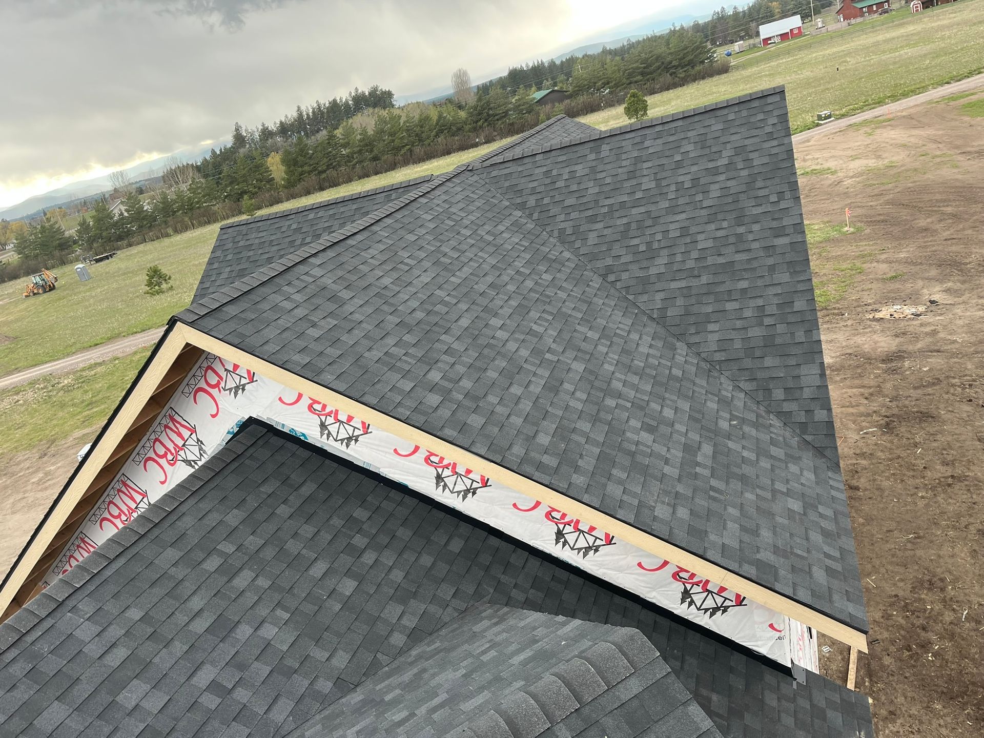 Dark gray shingled roof under construction with exposed ridge and a grassy landscape in the background