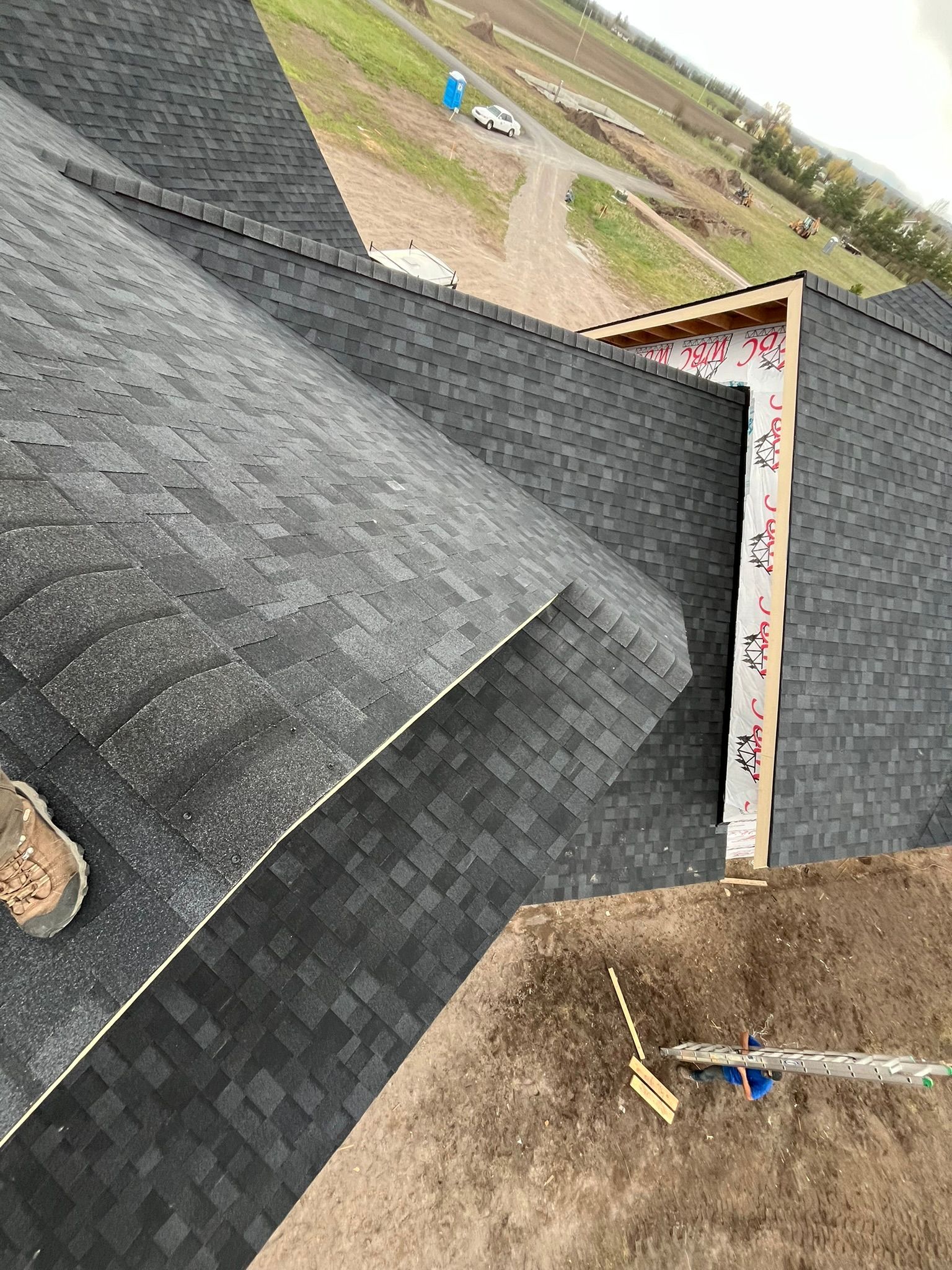 Roof under construction with dark shingles and a red-trimmed dormer against an overcast sky.