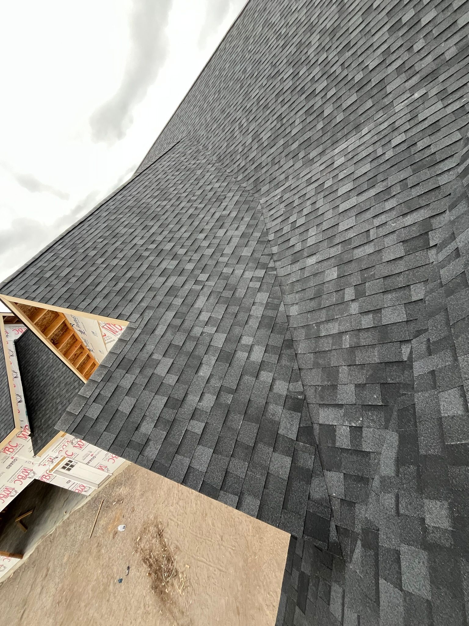 Steep gray shingled roof viewed from above with a small dormer opening and construction materials below