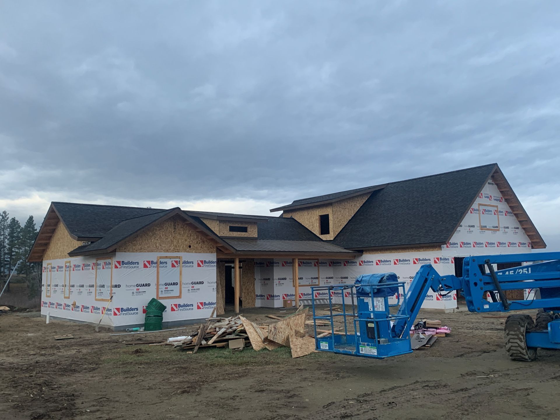 Partially built house under cloudy sky with construction wraps and blue boom lifts in front