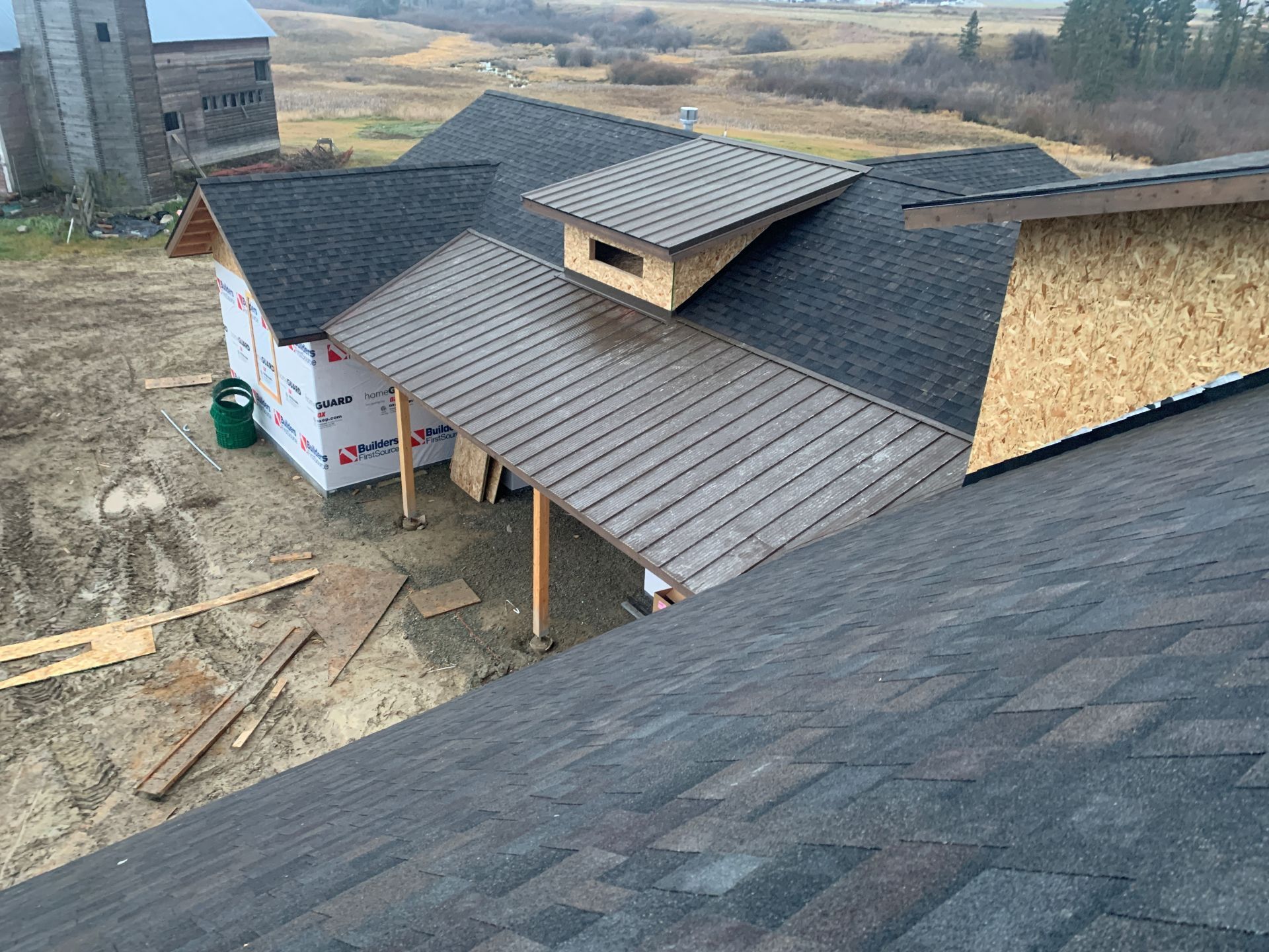 Roof under construction on a house with gray shingles, dormer window, and exposed plywood siding in a rural setting