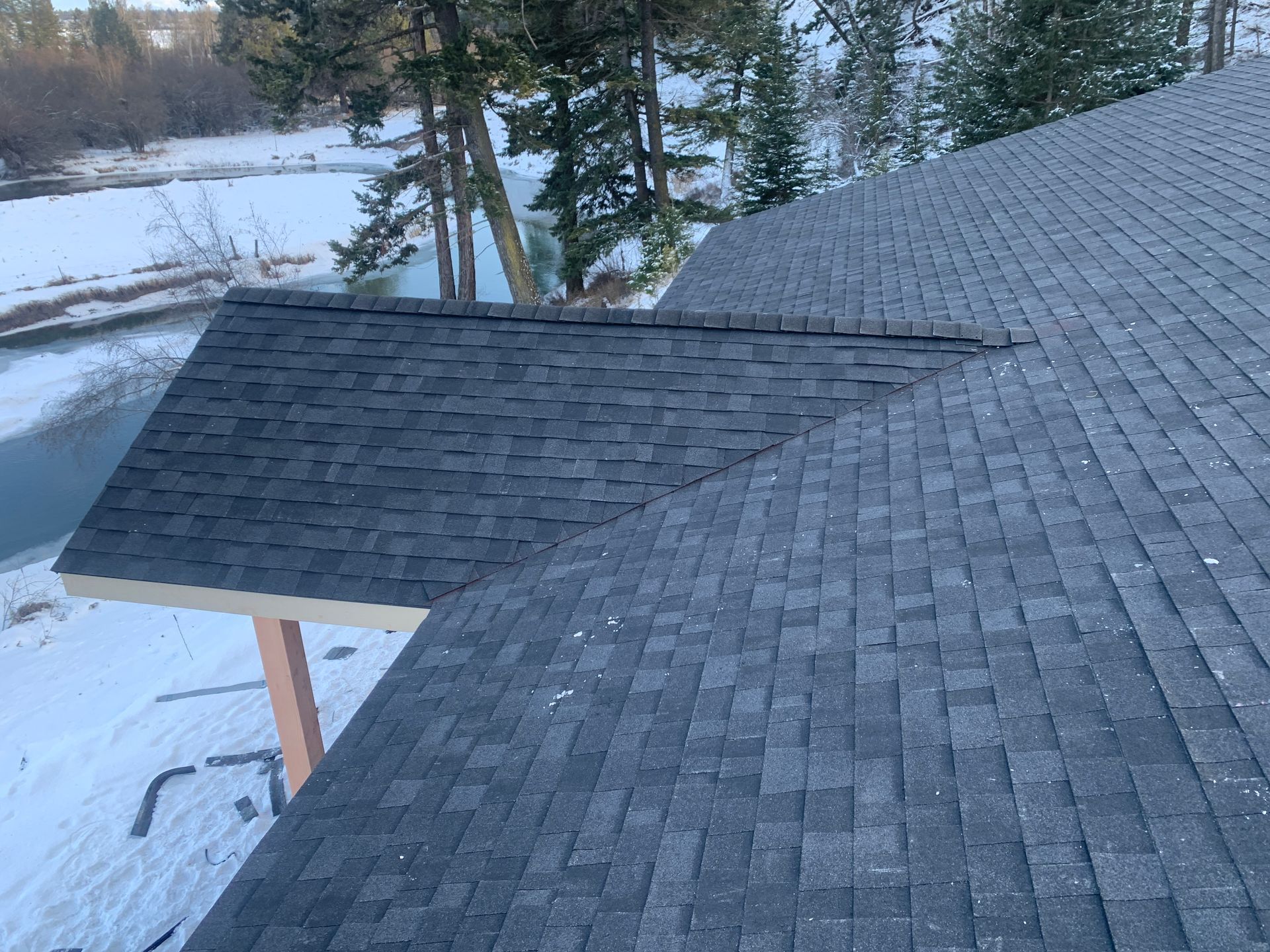 Snow-covered roof with dark shingles and a small dormer over a wooded winter scene