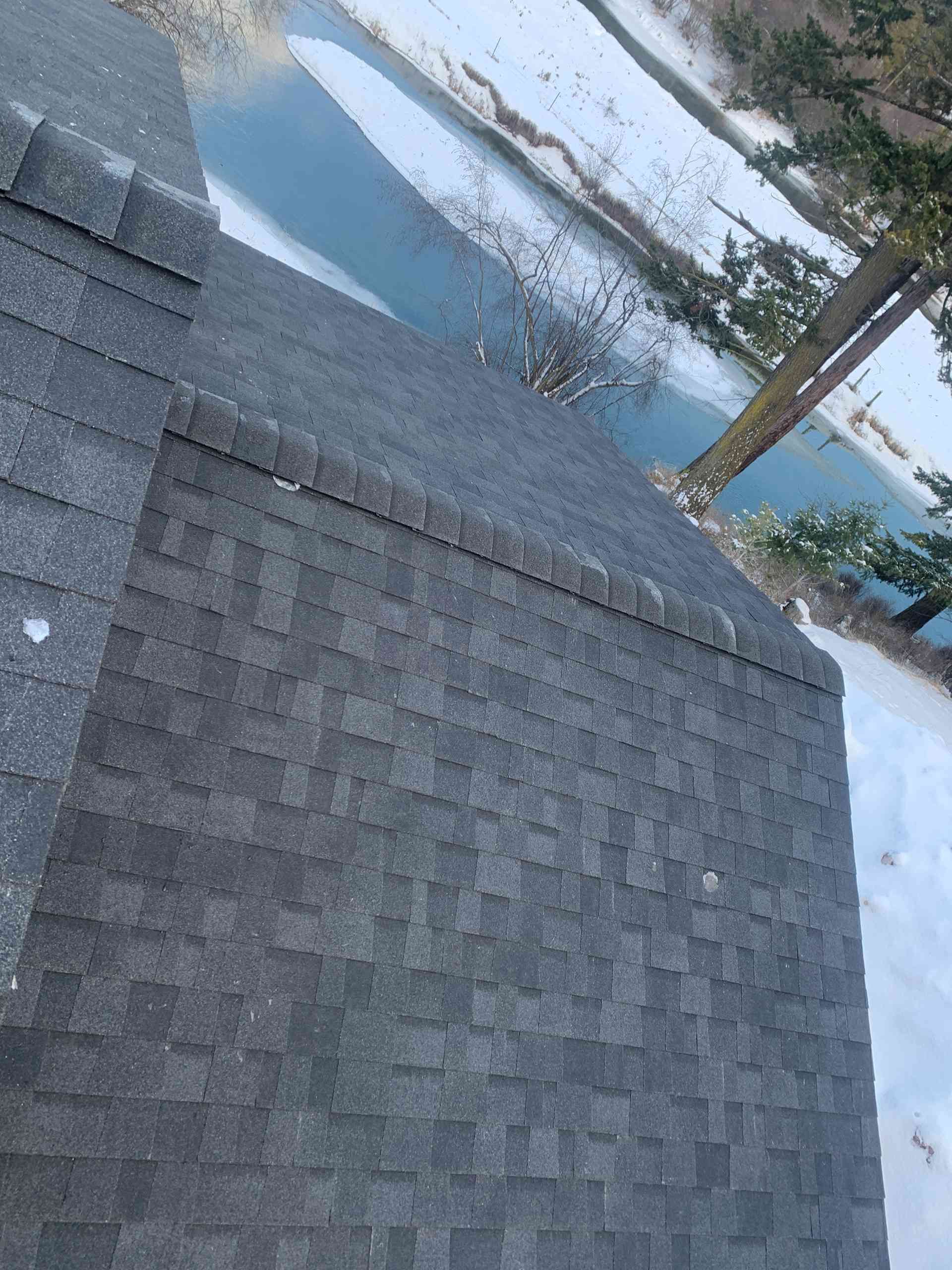 Snow-covered shingled roof beside a frozen pond with trees reflected in the water
