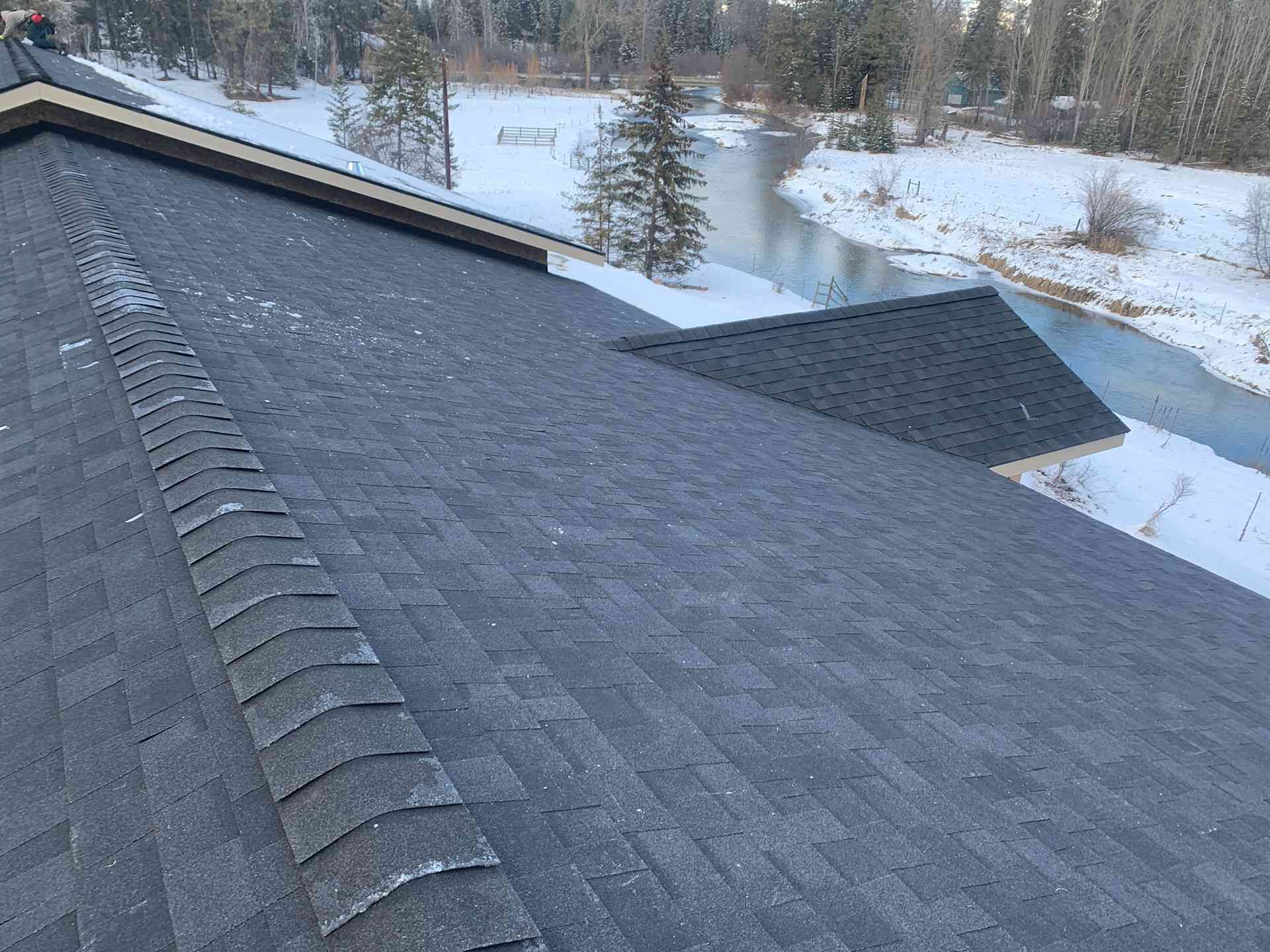 Snow-covered shingled roof with a dark ridge vent overlooking a snowy landscape and trees