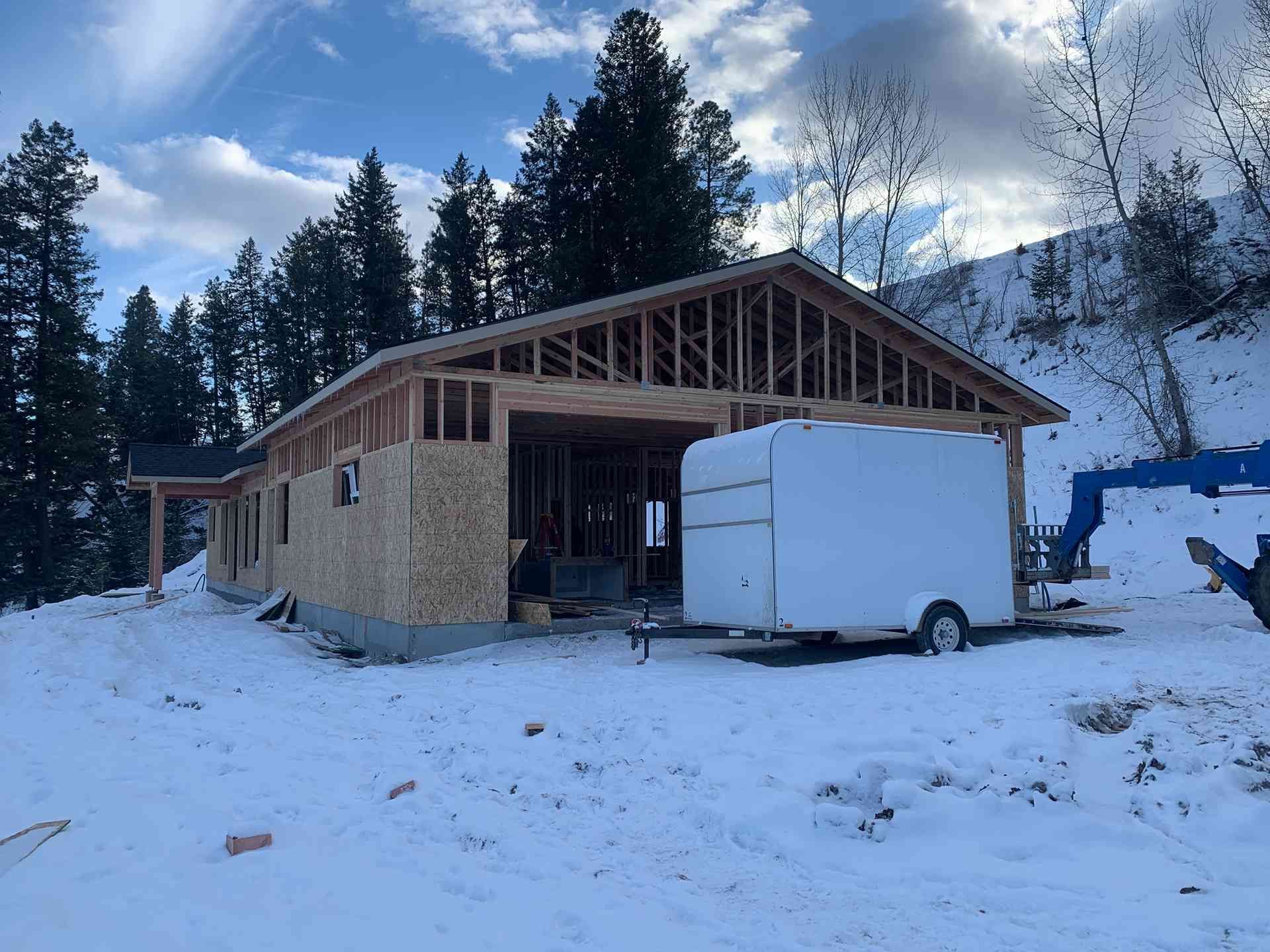 Snowy construction site with unfinished wooden house, white trailer, and pine trees in the background