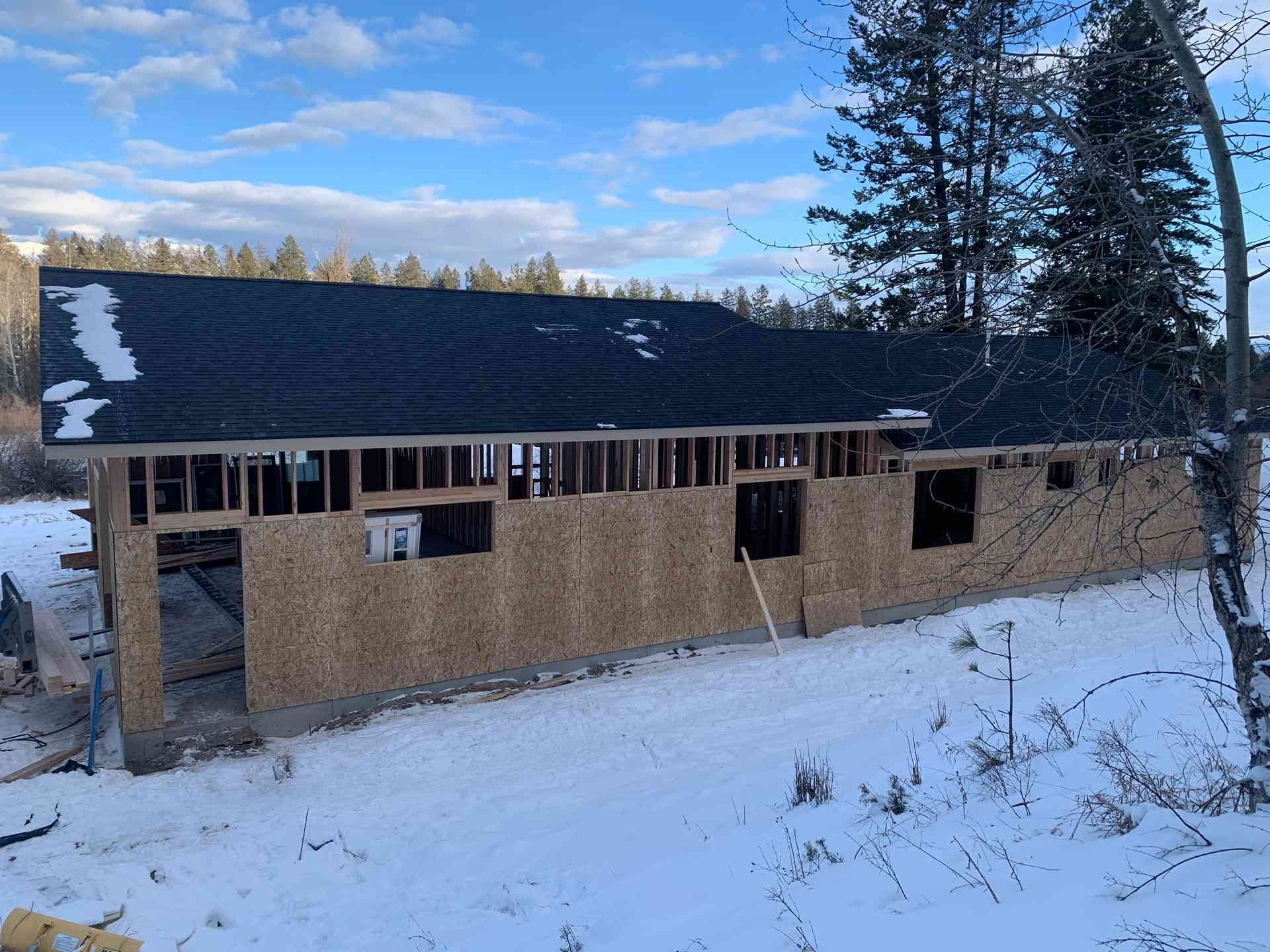 Snow-covered cabin under construction with unfinished walls and dark roof in a wooded winter setting