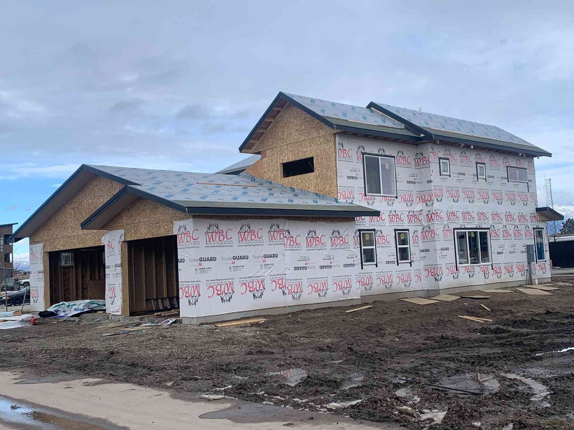 Partially built two-story house wrapped in insulation under a cloudy sky, with muddy ground around it