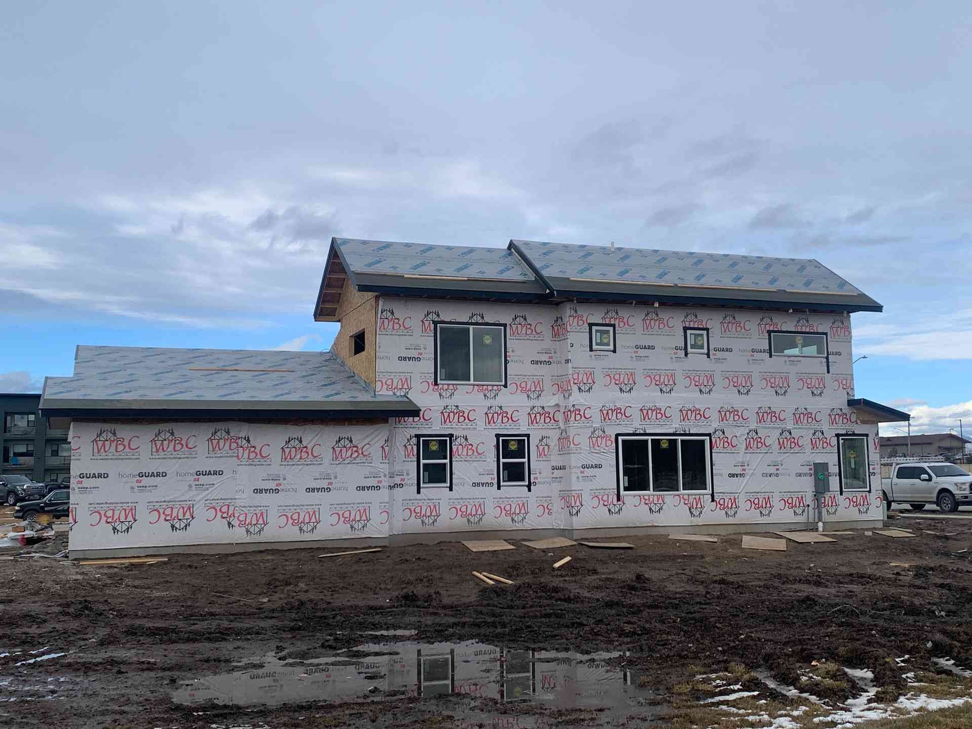 Partially built two-story house wrapped in white house wrap, set on a muddy construction site under a cloudy sky.