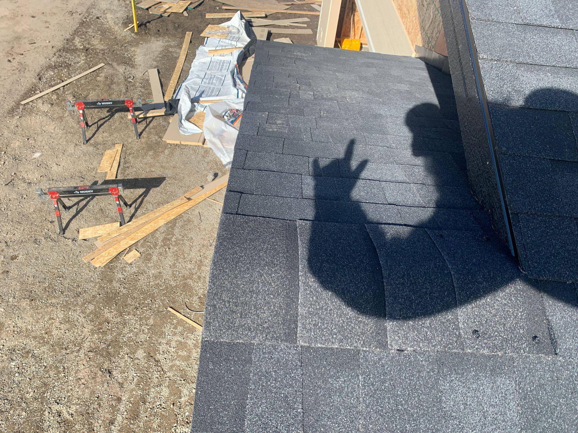 Roofing materials and tools on a roof edge with gray shingles and a shadow of the photographer's hand