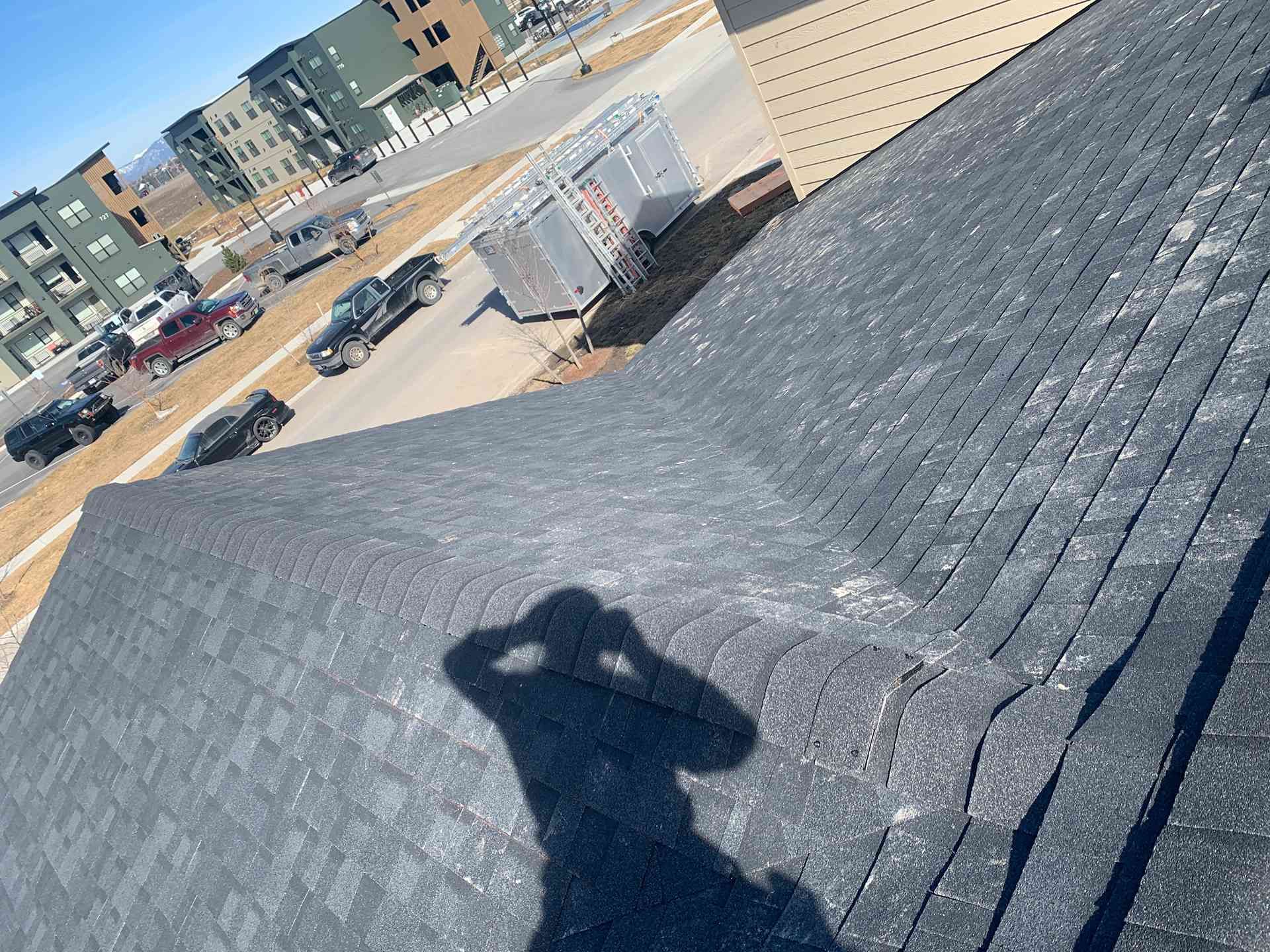 Tilted rooftop with dark shingles, a person’s shadow, and a parking lot with buildings below.