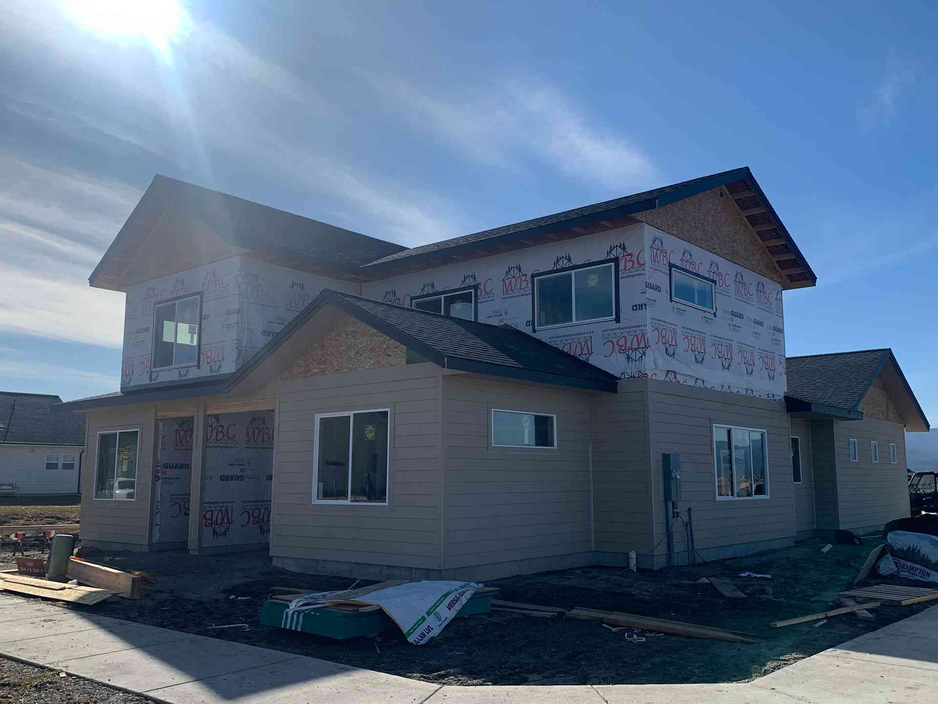 Two-story house under construction with siding, windows, and roof in a sunny open lot