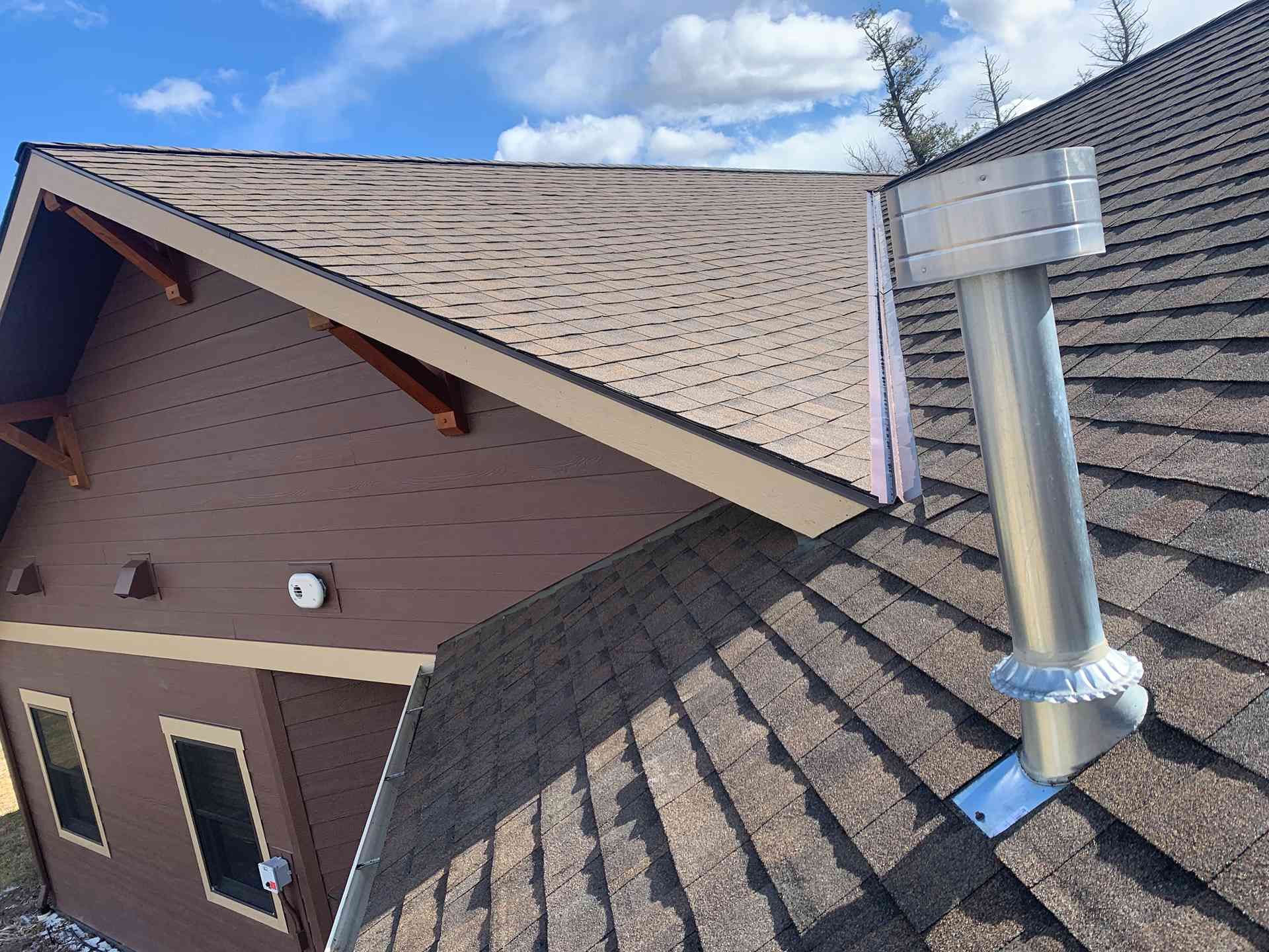 Roof with gray shingles and a silver vent pipe beside a brown house under a blue sky