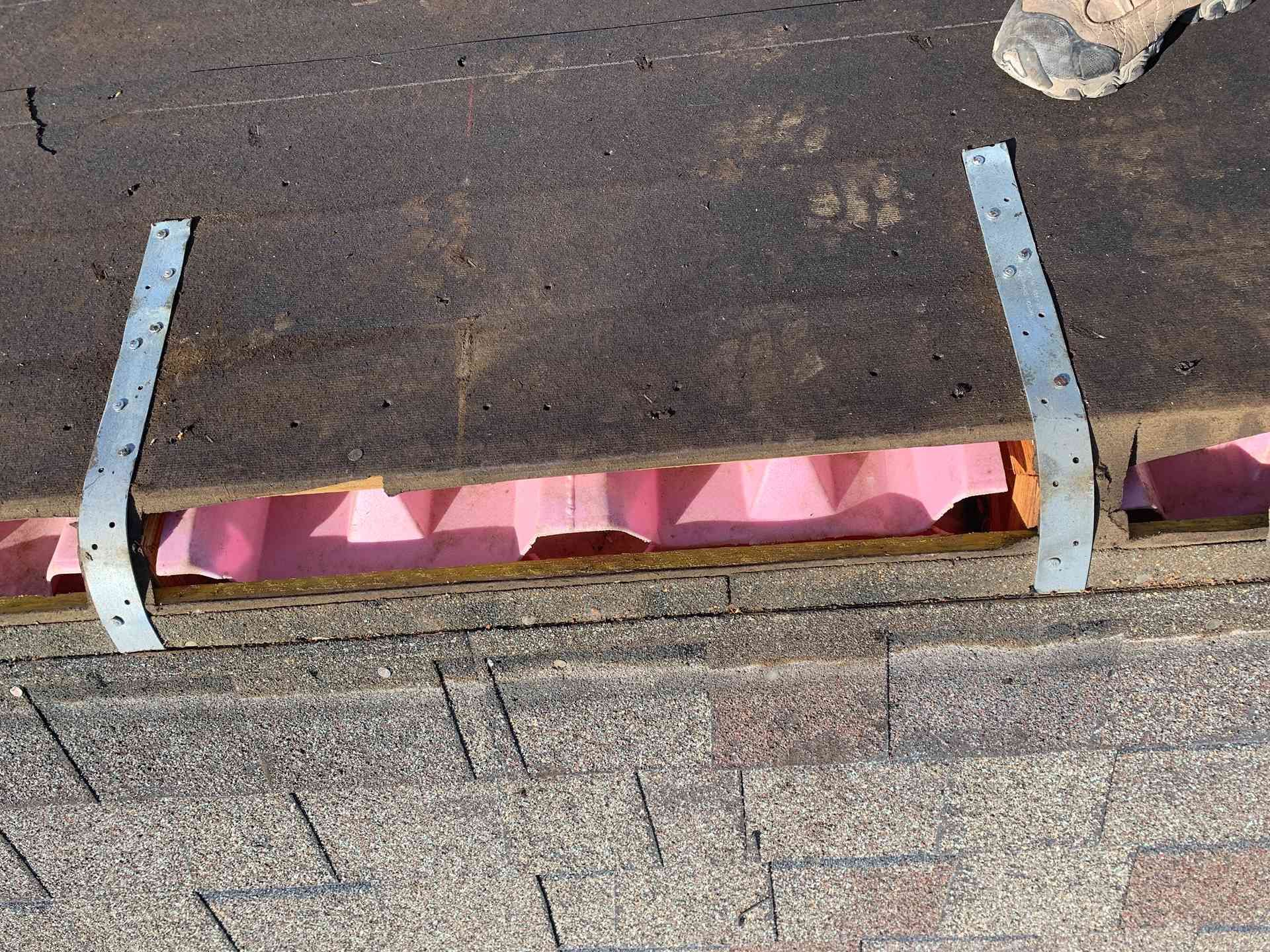 Close-up of a roof edge with two gray metal brackets and pink flashing beneath shingles