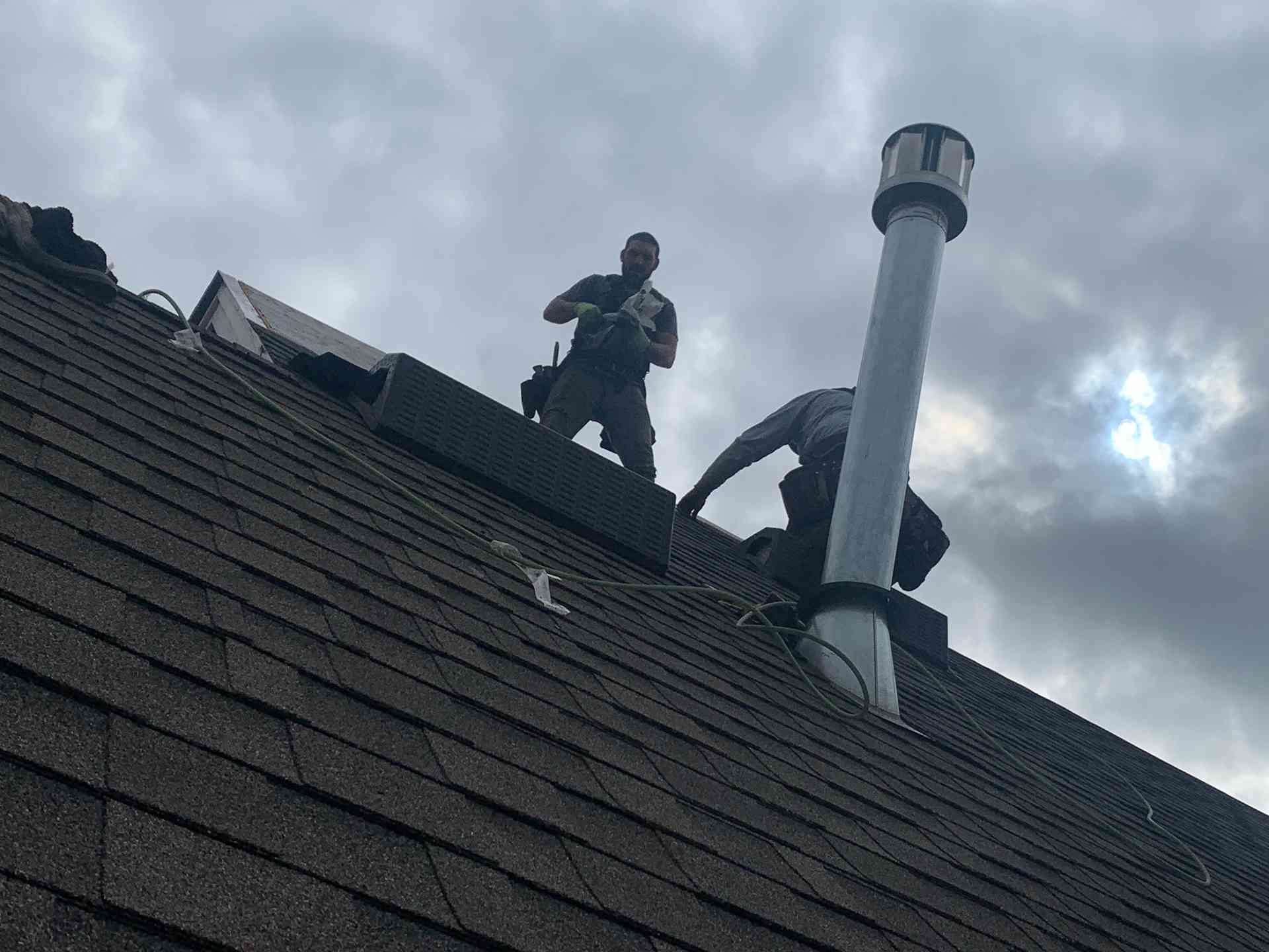 Person standing on a shingled roof beside a metal vent pipe under cloudy sky