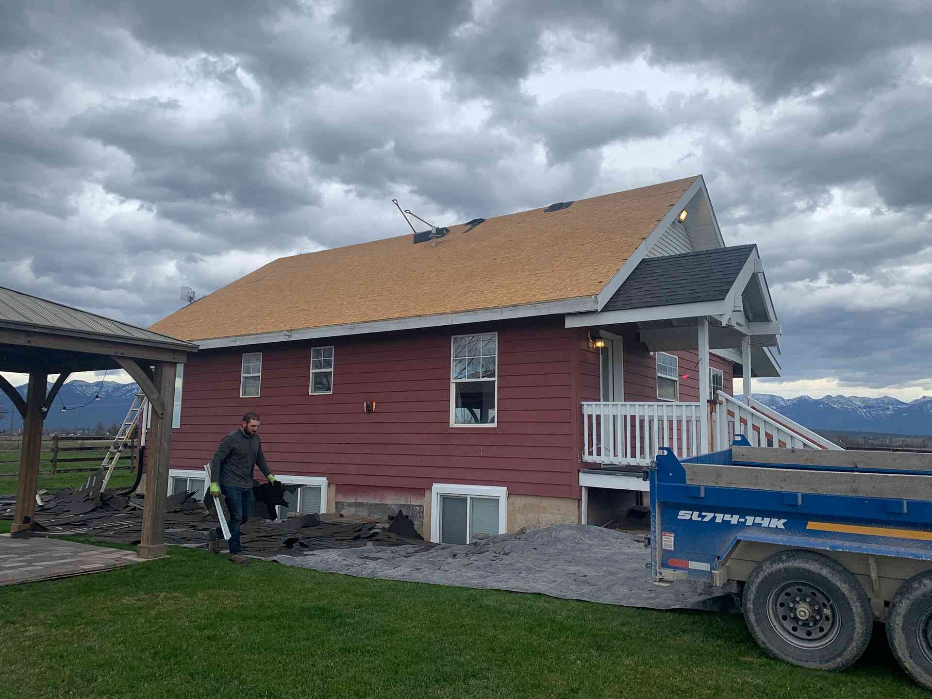 Red house with tan roof on a gravel lot, mountains in the background, and a blue trailer parked beside it