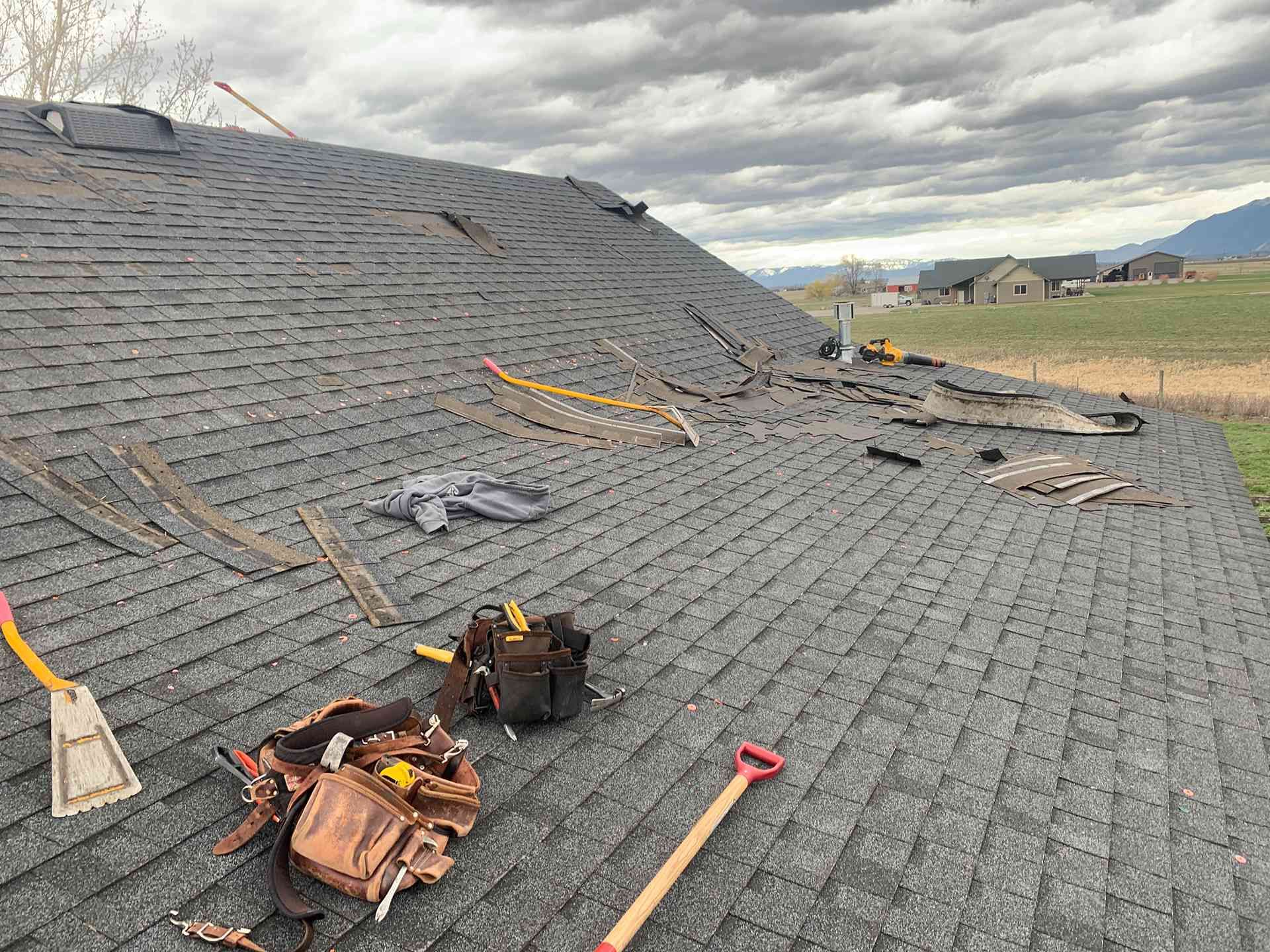 Roof under repair with gray shingles, scattered tools, and cloudy sky beyond the ridge