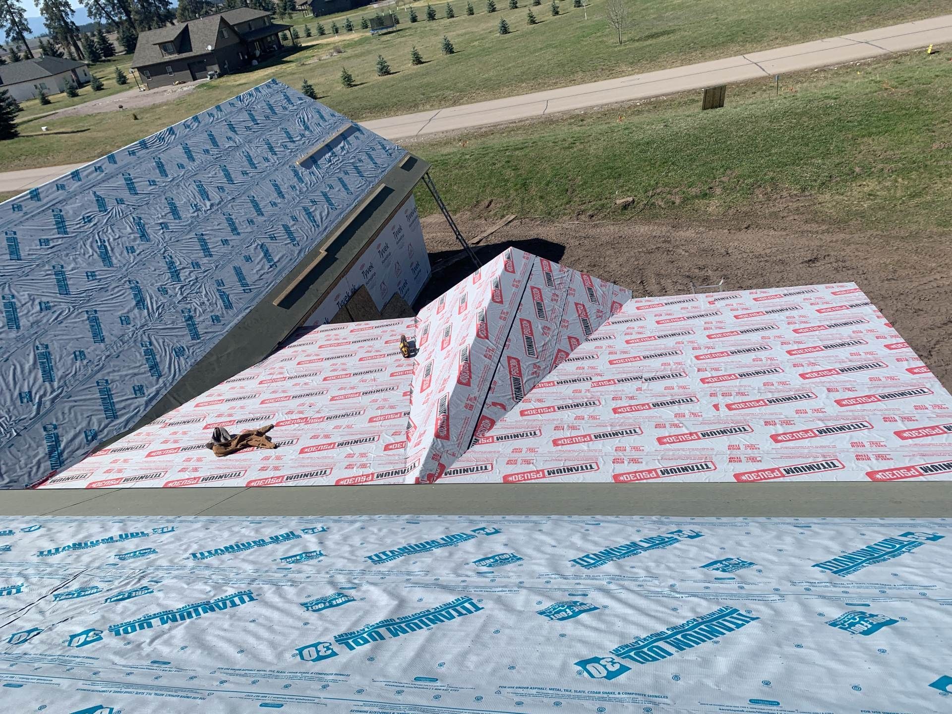 Stacks of wrapped roofing shingles on a grassy rooftop, with blue and white packaging visible.