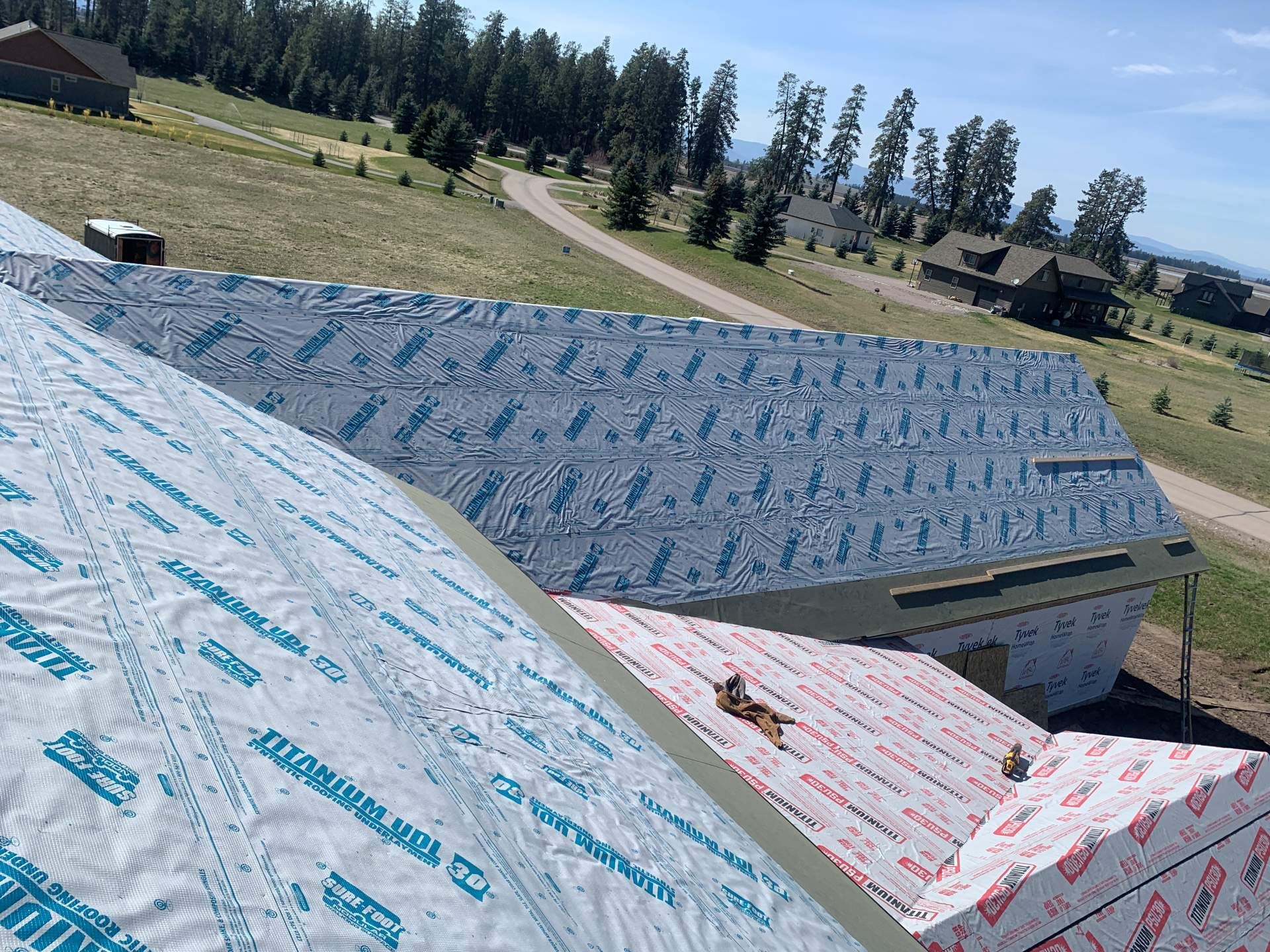 Roof under construction with blue and pink house wrap in a rural hillside setting