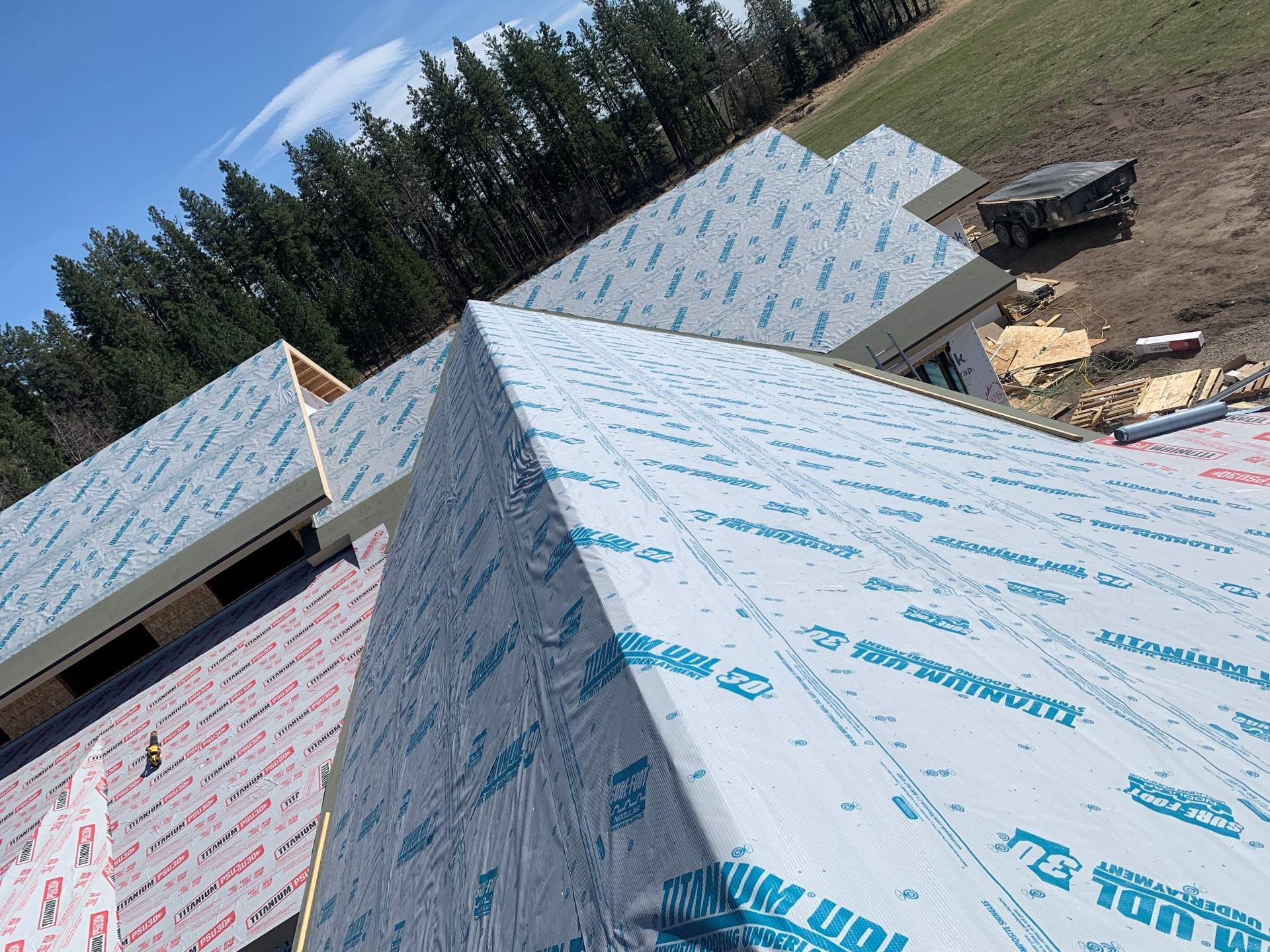 Roof under construction with blue-white protective wrap on steep gables and trees in the background