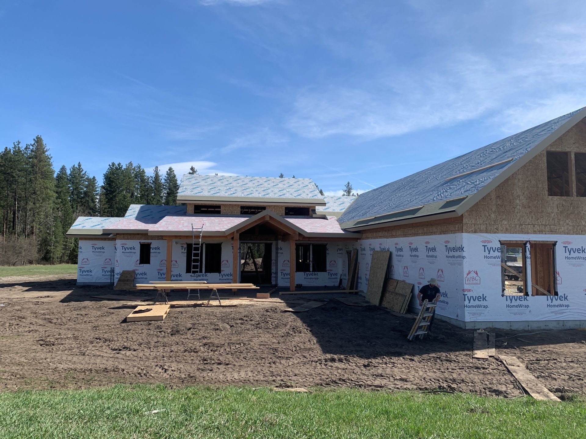 Partially built house with exposed framing, white wrap, and a dirt yard under a blue sky