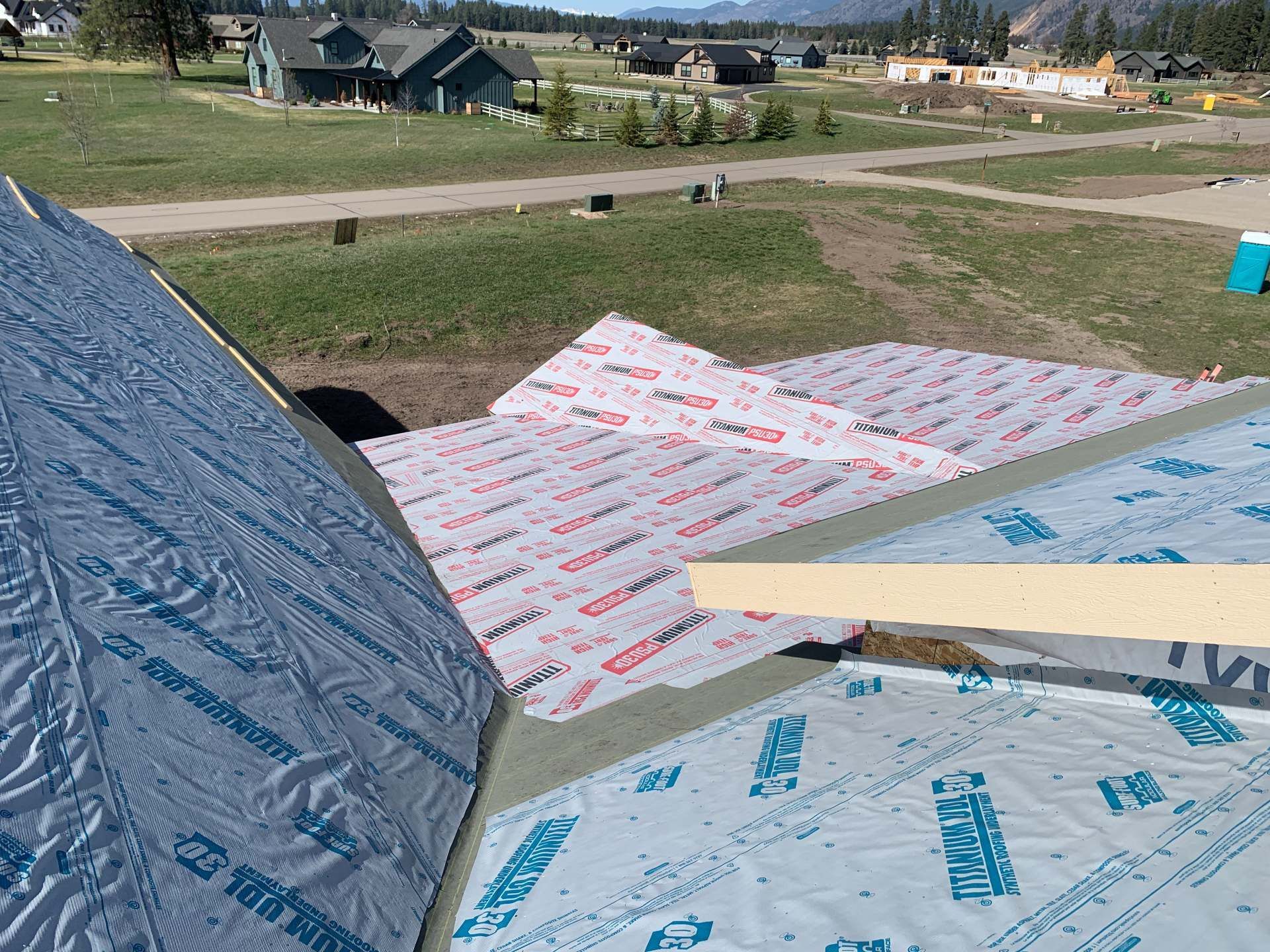 House roof under construction with blue and white roofing underlayment on a sloped roof, grassy neighborhood beyond