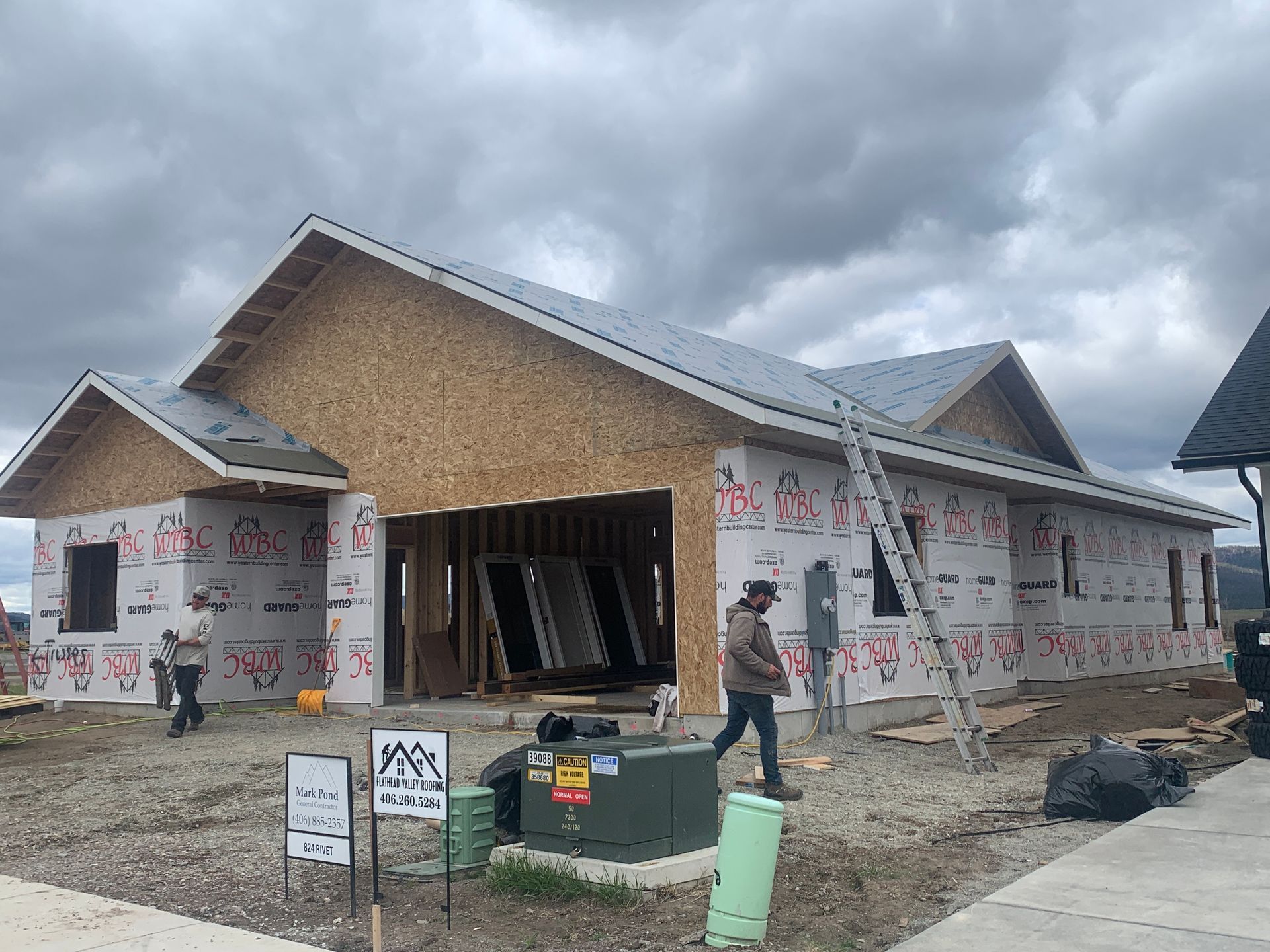Partly built house with exposed framing, workers, and stormy sky on a muddy construction site