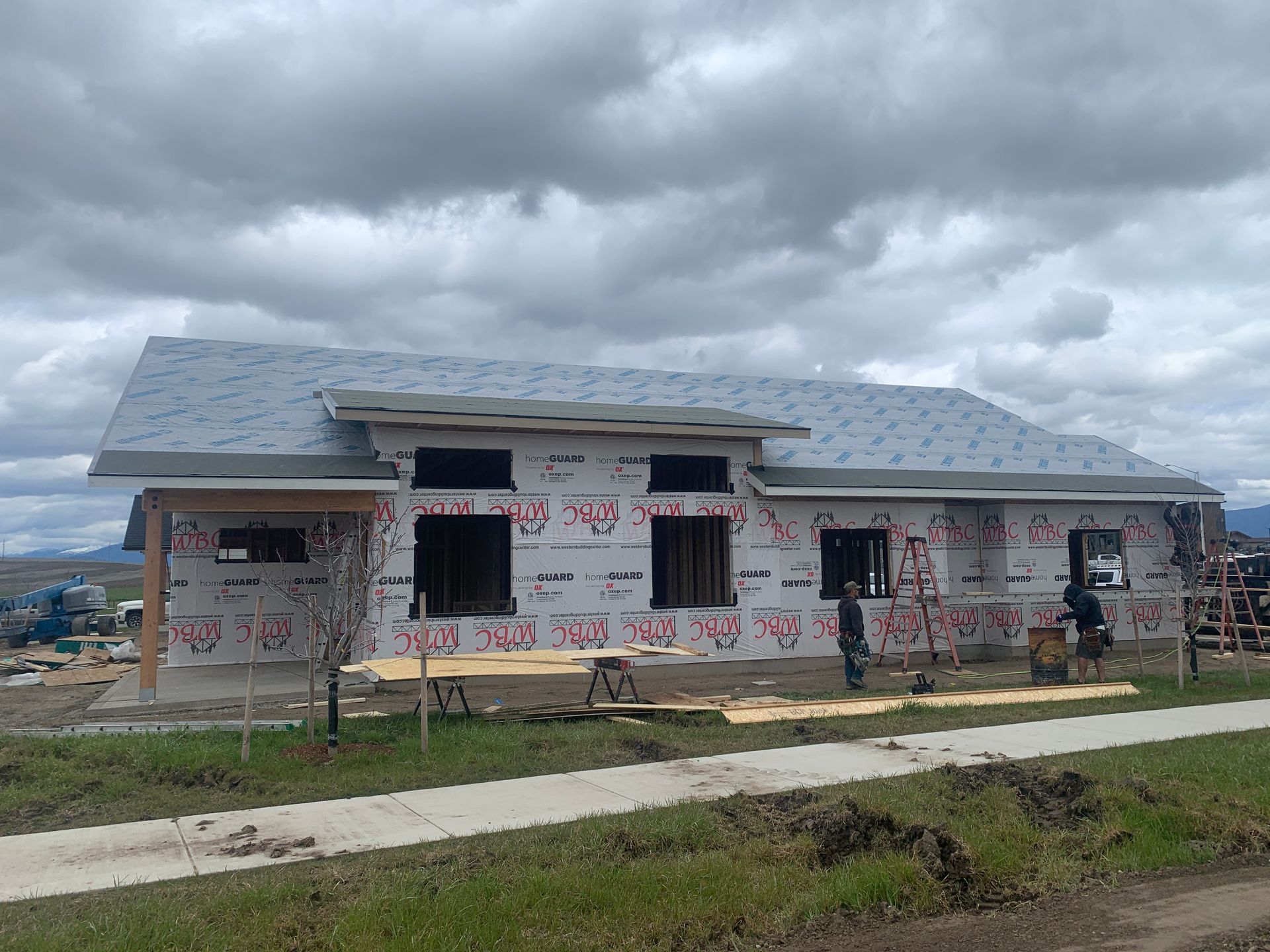 Partially built house with wrapped exterior under a cloudy sky, viewed from across a grassy lot.