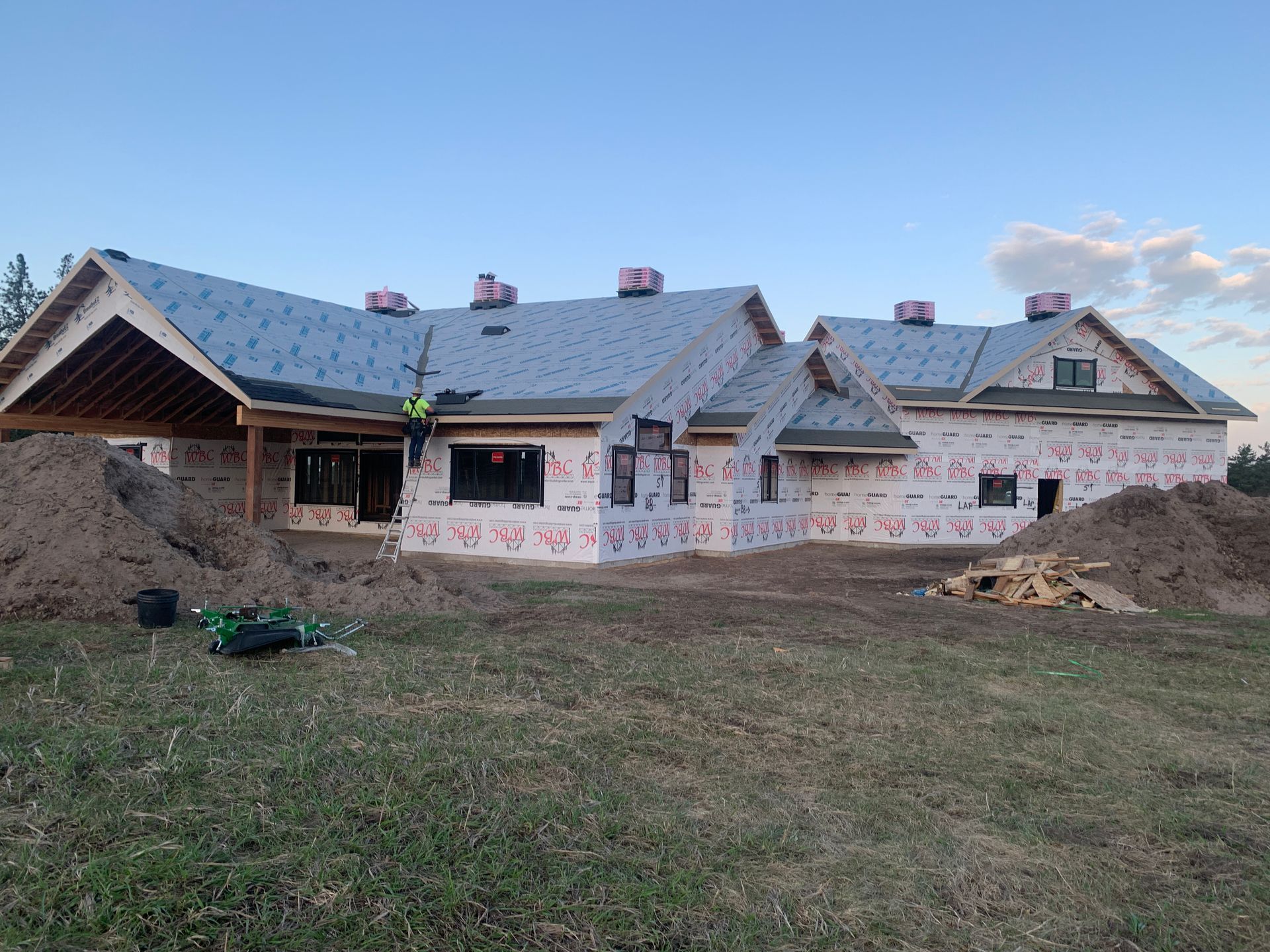 A house under construction with a gray roof, exposed walls, and piles of dirt in front under a blue sky