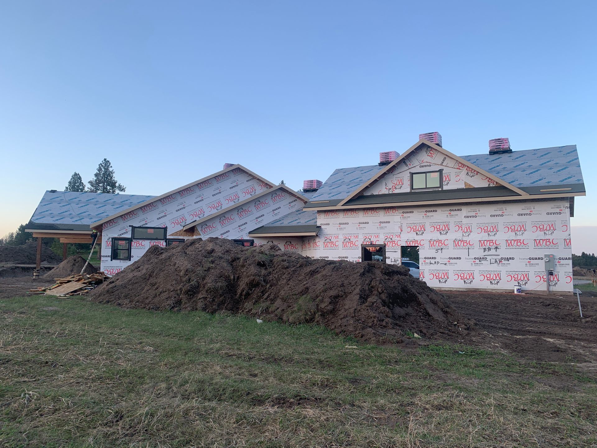 Partially built house with white wrap and gray roof beside a dirt mound under a blue sky