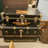 Stacked black vintage trunks with brass hardware in a shop corner, one with a winged emblem and keyhole.