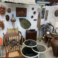 Eclectic antique shop display with chairs, woven baskets, wall decor, and a glass-top table
