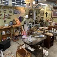 Cluttered antique shop interior with shelves of vintage items, framed art, and a central table of assorted goods