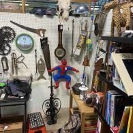 A cluttered shop display with hanging tools, clocks, books, and a Spider-Man doll in the center.