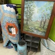 Framed landscape painting beside a quilted textile and small lantern in a thrift-store display