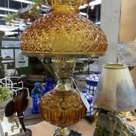 Ornate amber glass table lamp with a woven shade displayed in a shop, beside a smaller lamp.